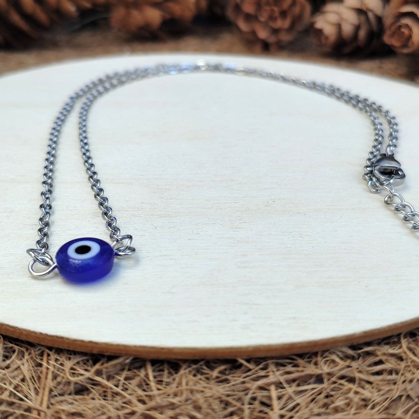 Necklace with a blue evil eye charm on a white plate surrounded by pine cones.