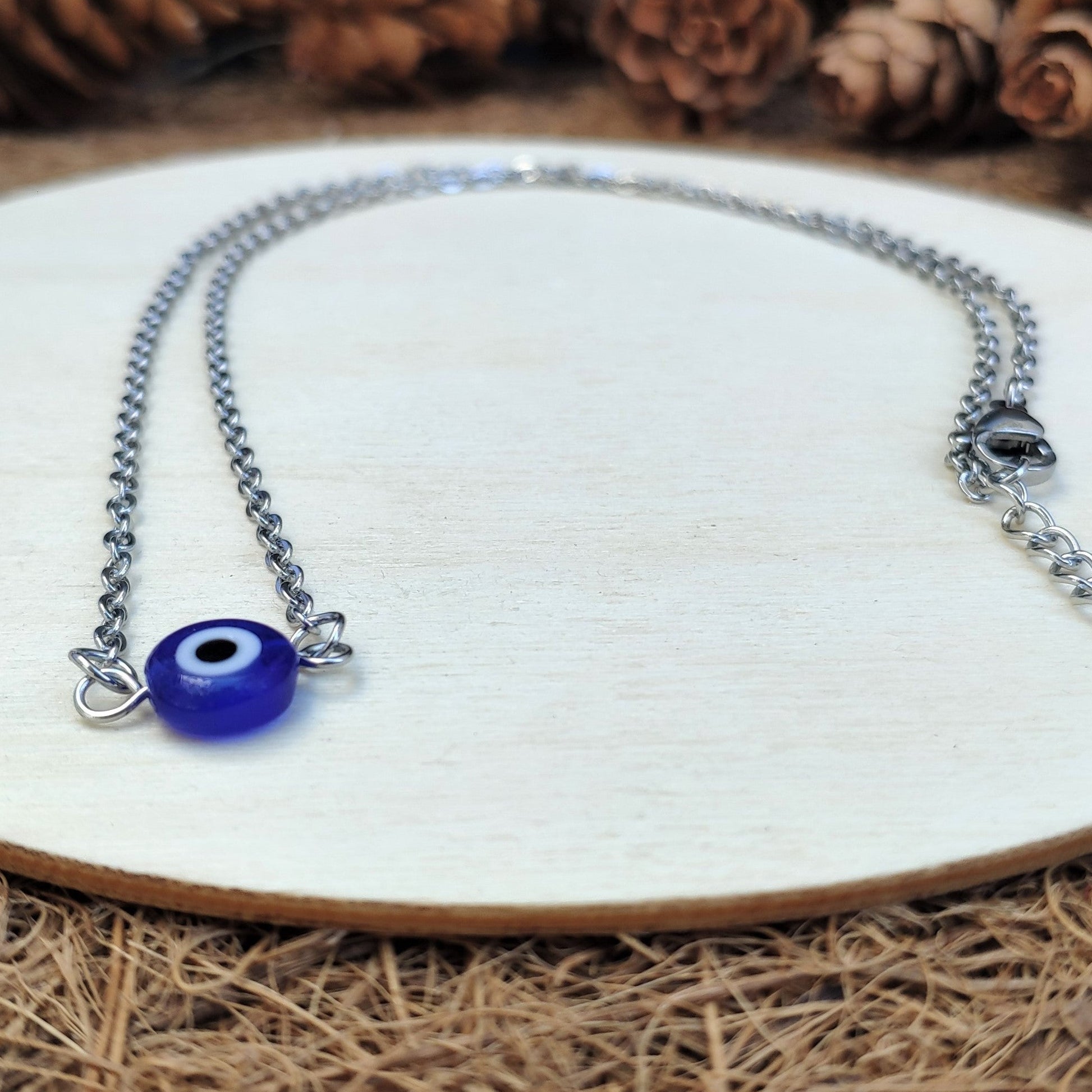 Necklace with a blue evil eye charm on a white plate surrounded by pine cones.