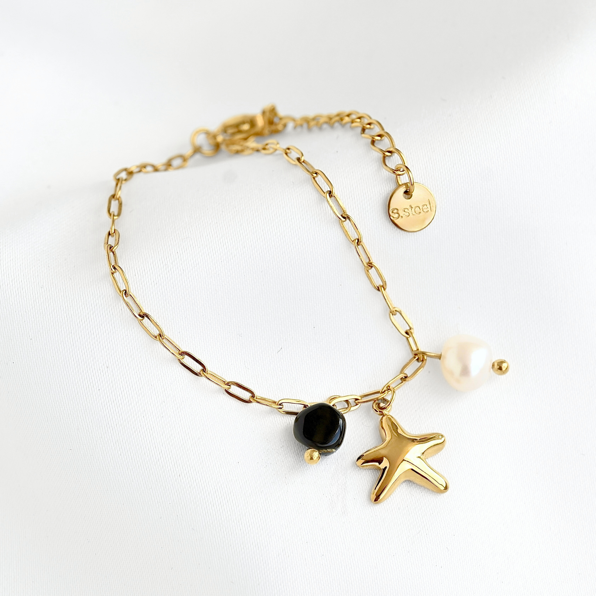 A gold chain bracelet with a lobster clasp, featuring dangling charms including a starfish, a black bead, a white pearl, and a round gold tag engraved with 'S.steel'.