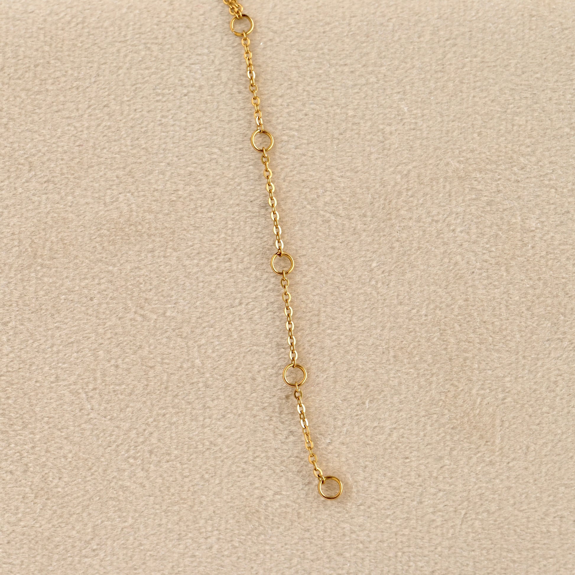 A close-up of a delicate gold chain extender with four small circular links and a drop loop, laid on a textured beige surface.