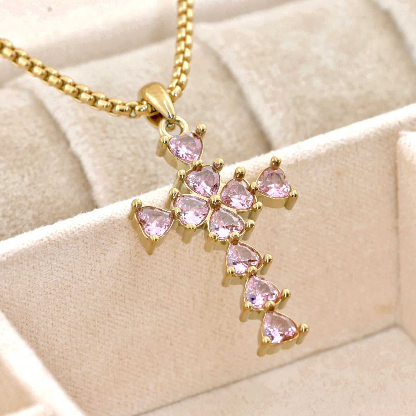 Gold cross necklace with pink heart shaped diamonds on a beige fabric background