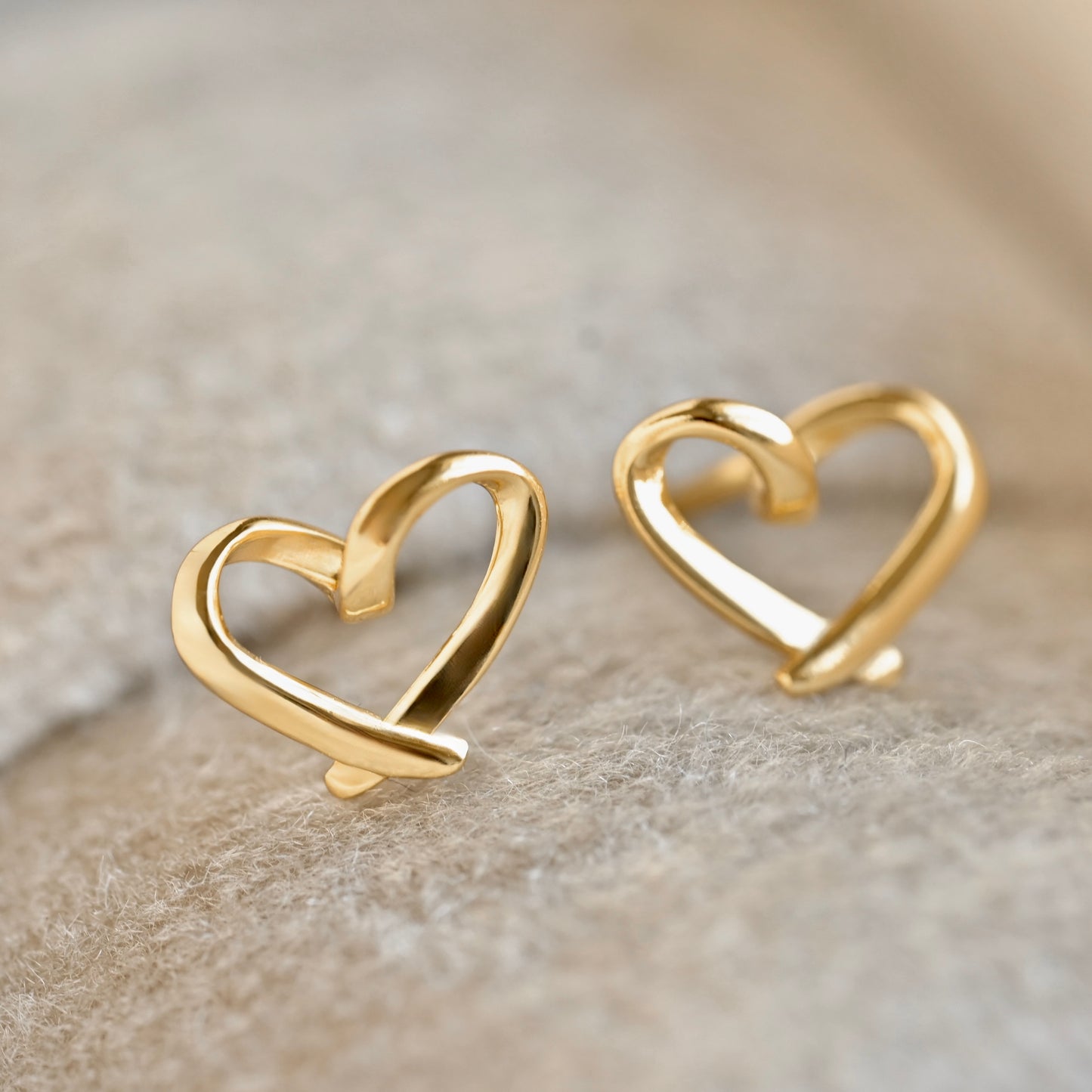 A pair of gold heart-shaped stud earrings with a polished finish, resting on a textured beige surface.