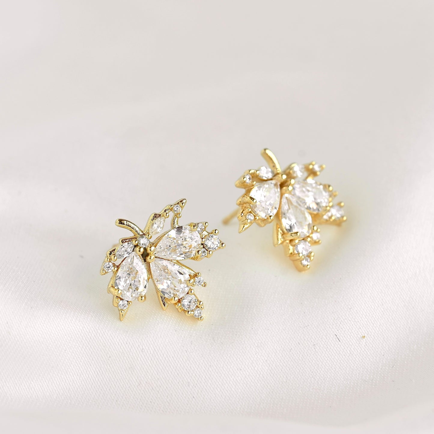 A pair of gold leaf-shaped stud earrings adorned with clear marquise and round crystals, displayed on a soft white fabric background.