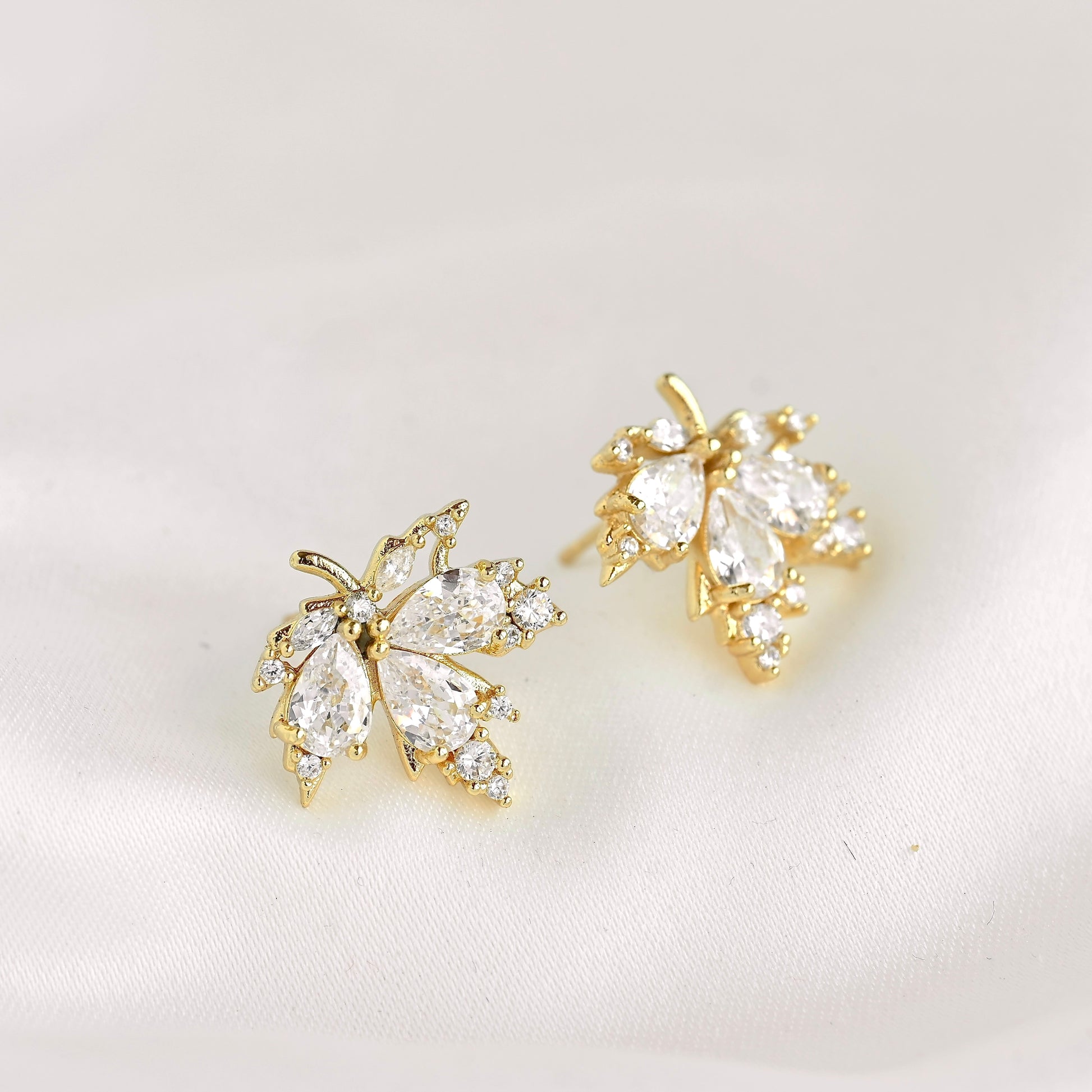 A pair of gold leaf-shaped stud earrings adorned with clear marquise and round crystals, displayed on a soft white fabric background.