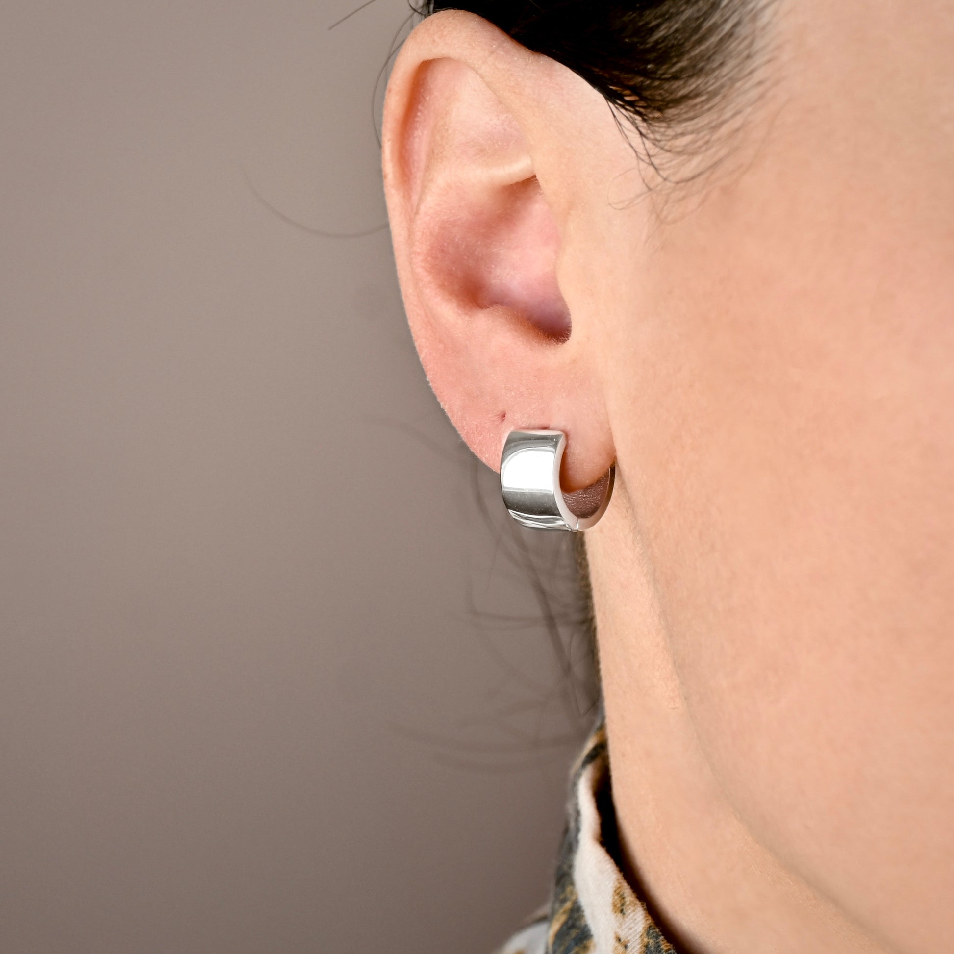A close-up of a person's ear wearing  polished silver wide and thick hoop earrings against a plain taupe background.