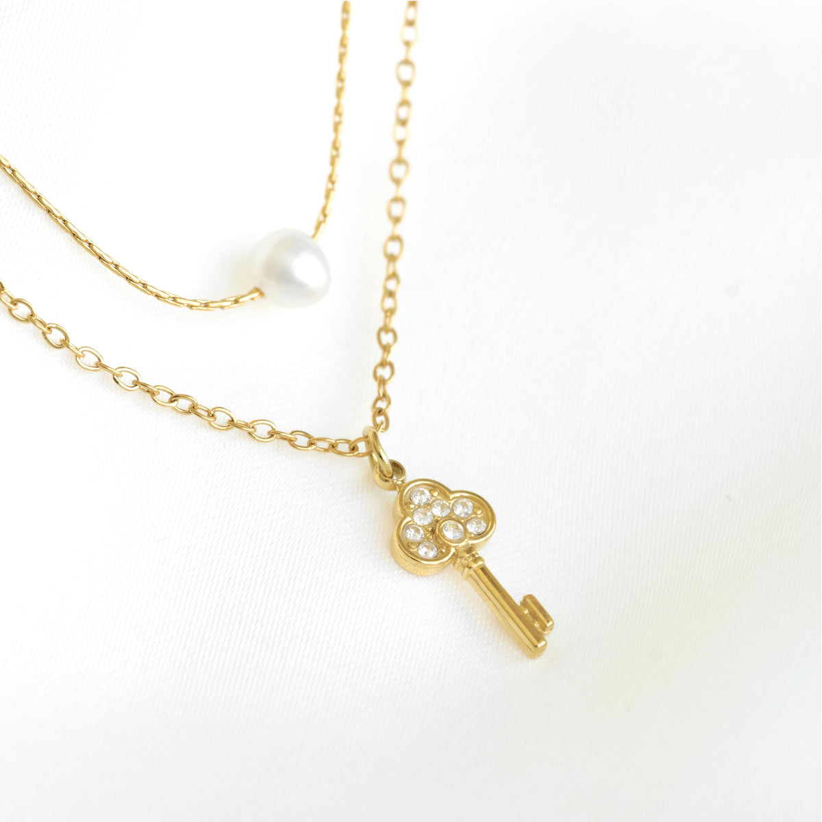 A gold-tone layered necklace featuring a key-shaped pendant adorned with small clear stones and a single white pearl bead on a delicate chain, displayed on a clean white background.