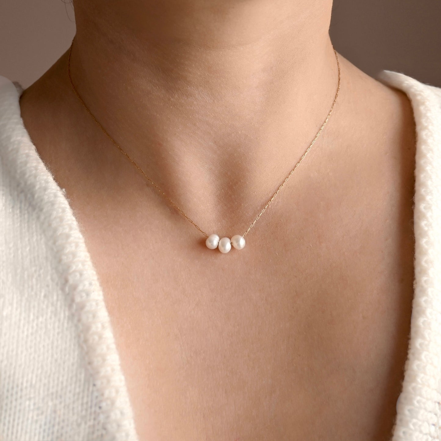 An ultra thin gold paperclip chain necklace with three small white pearls, worn on a model's neck over a white textured top.