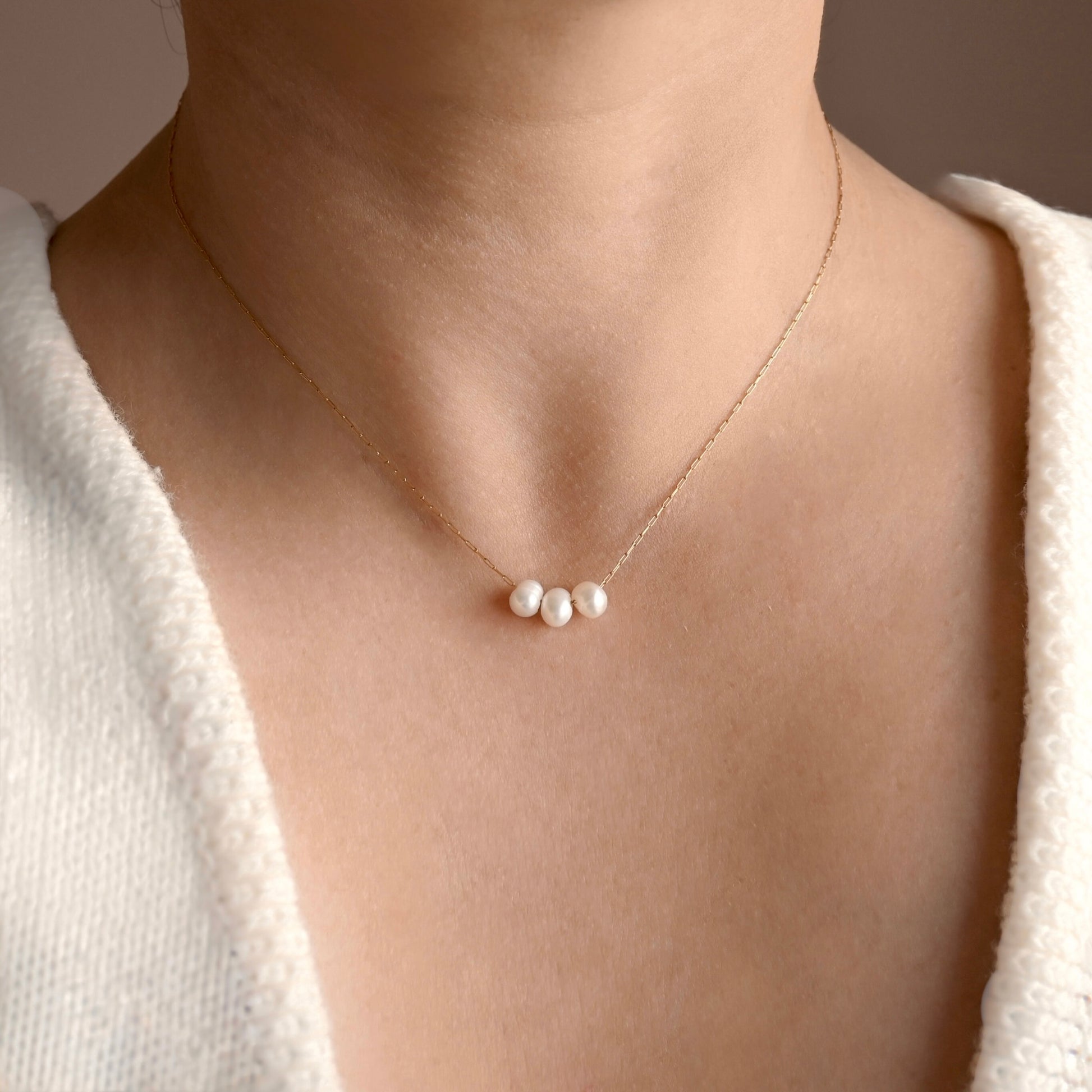 An ultra thin gold paperclip chain necklace with three small white pearls, worn on a model's neck over a white textured top.
