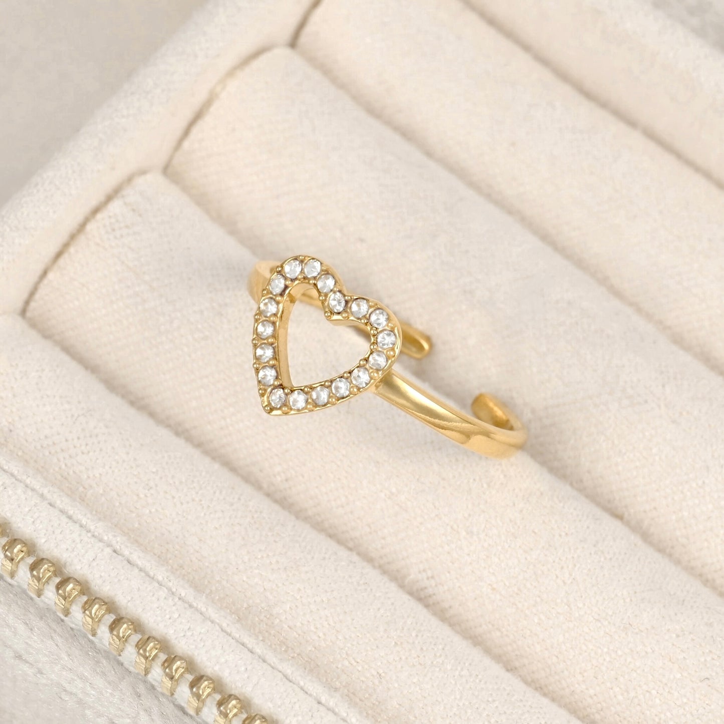 A gold adjustable ring with an open heart design set with small clear stones, displayed on a cream-colored jewelry box.