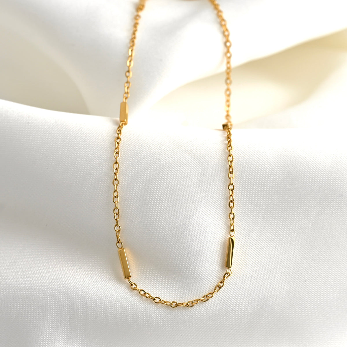 A delicate gold chain necklace with four small polished gold bar accents, displayed on a soft white satin fabric background.