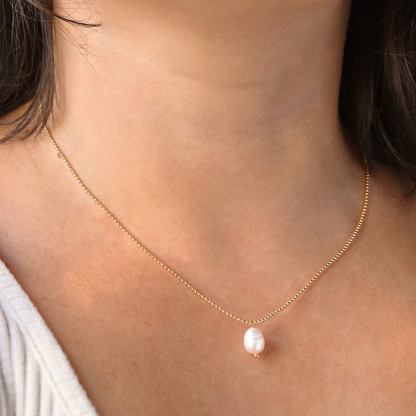 A delicate gold chain necklace with a single white baroque pearl pendant, shown worn on a model's neck.
