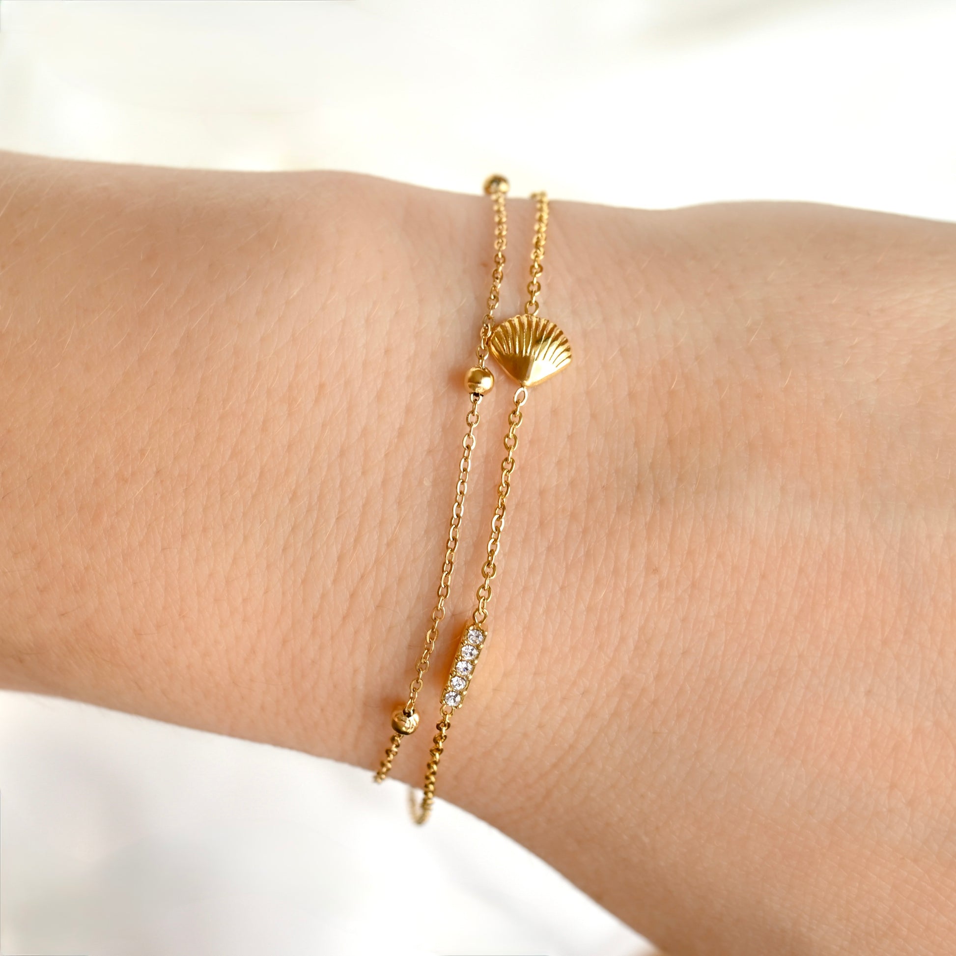 A delicate gold double-strand bracelet with a seashell charm and a small bar of clear stones, shown worn on a person's wrist.