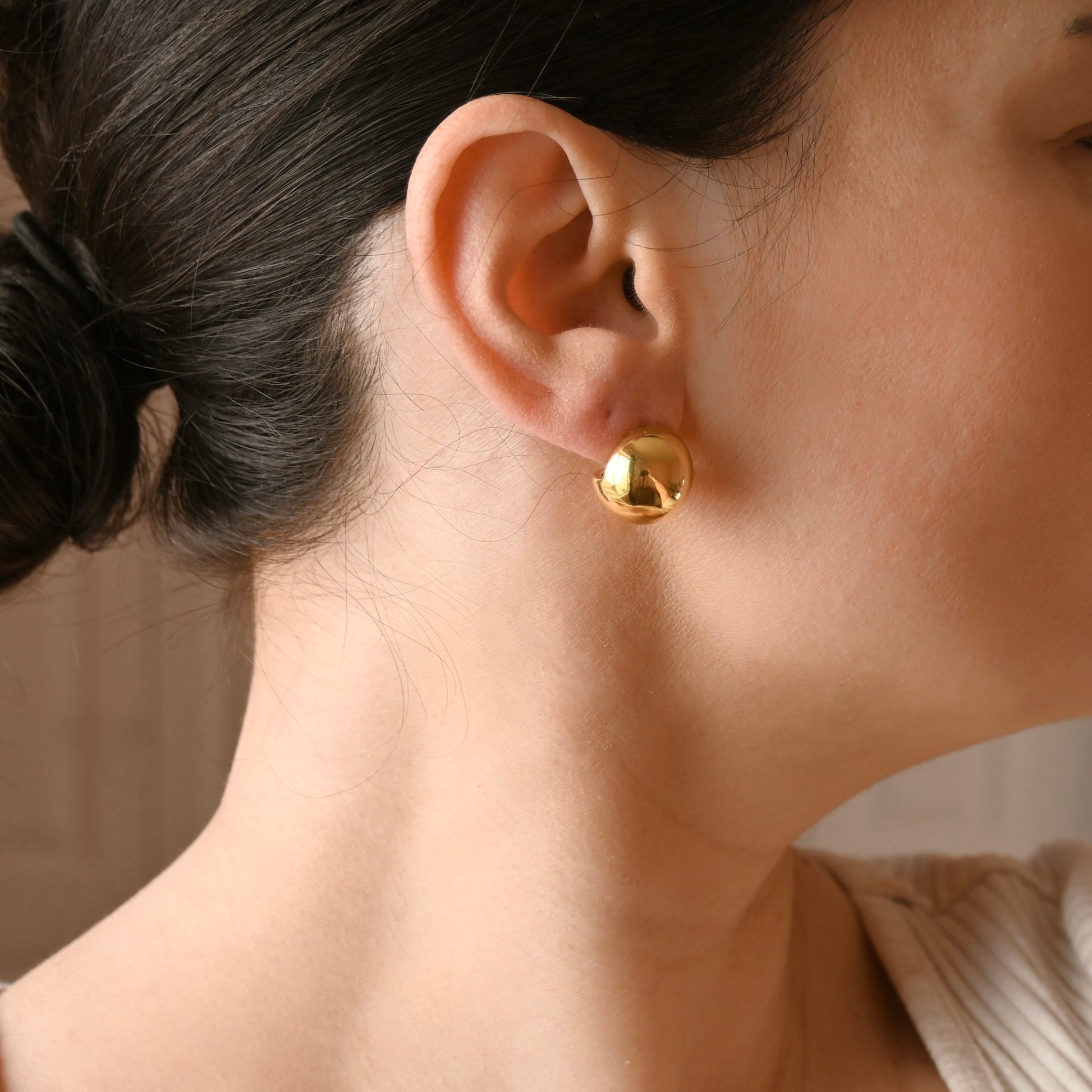 A close-up of a person's ear wearing a small, polished gold sphere stud earring.