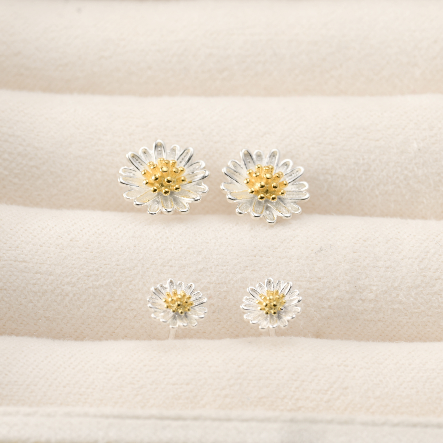 A pair of small and medium silver daisy earrings on a beige cushion