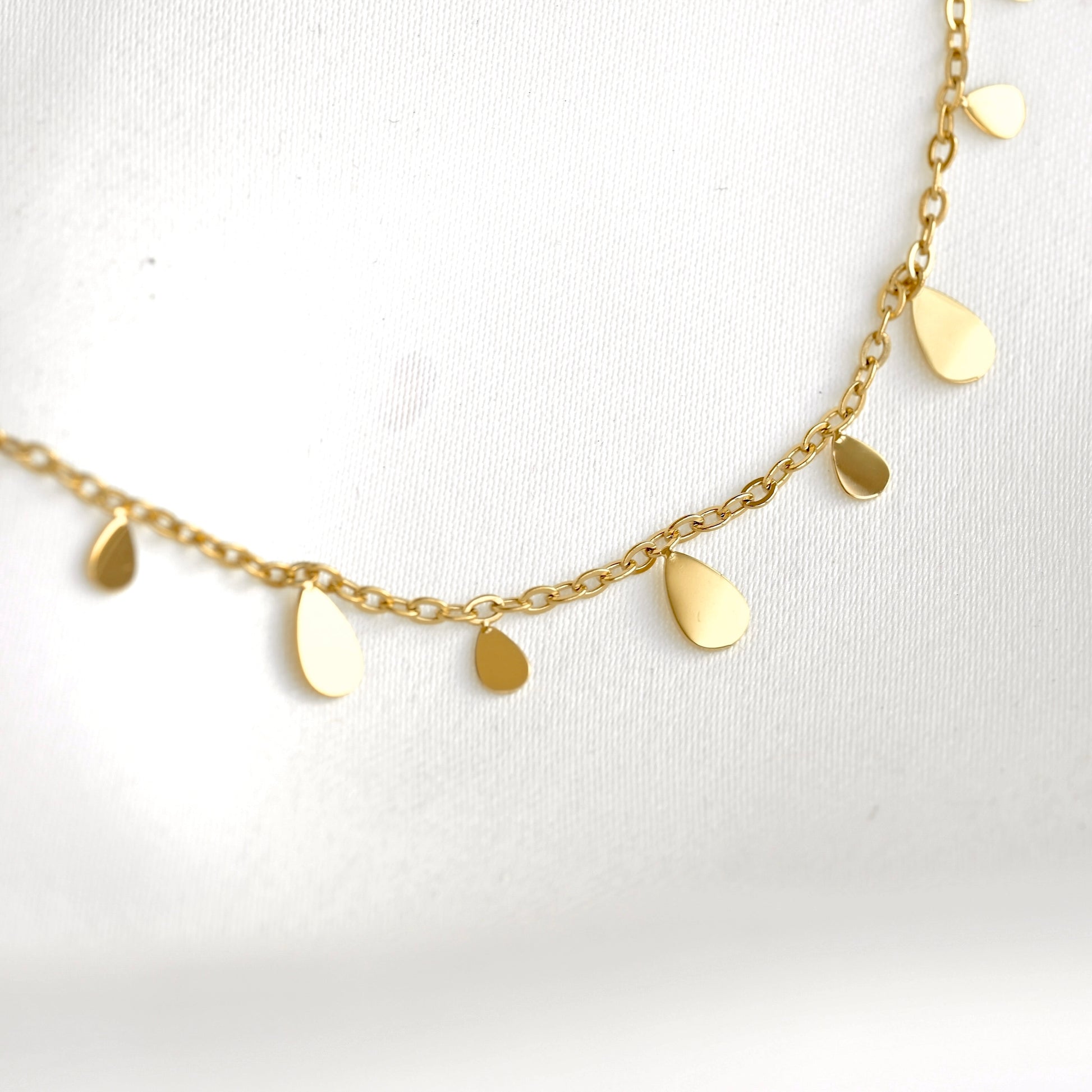 A delicate gold chain necklace featuring small, flat teardrop-shaped metal charms, displayed on a textured white fabric background.