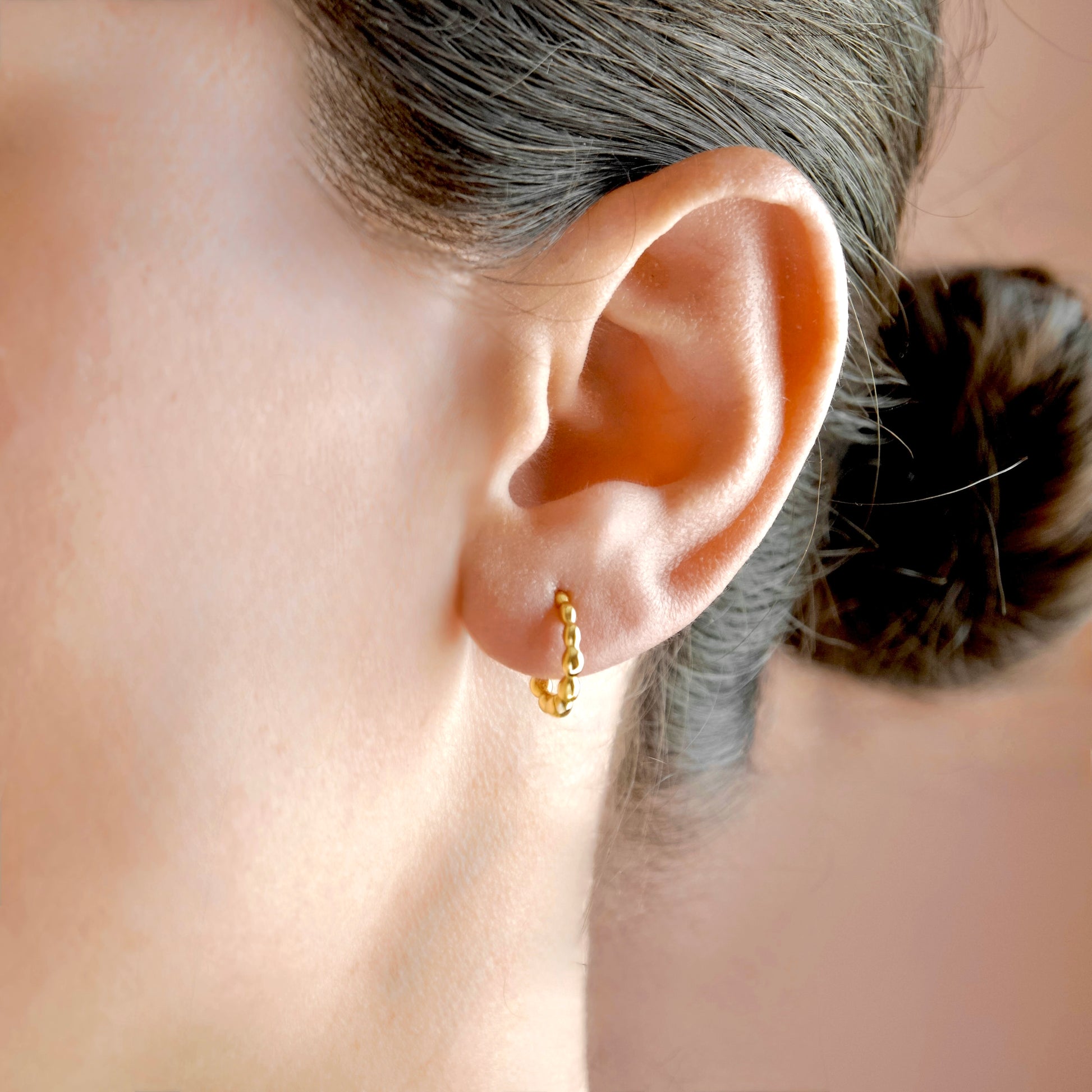 A close-up of a person's ear wearing a small gold huggie hoop earring, with the hair pulled back into a bun against a soft peach-toned background.