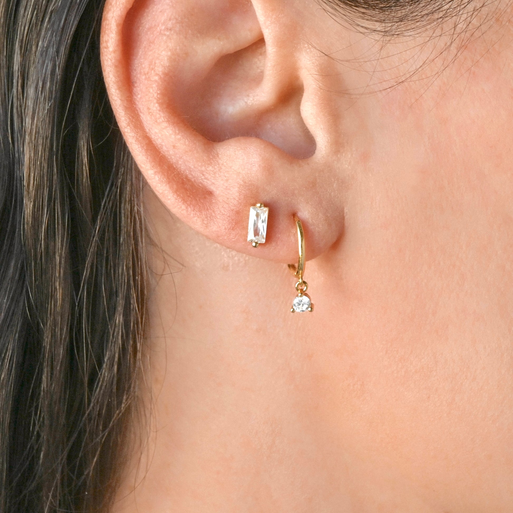 Close-up of a person wearing gold hoop earrings with clear gemstones and a rectangular diamond bar stud in the second hole.