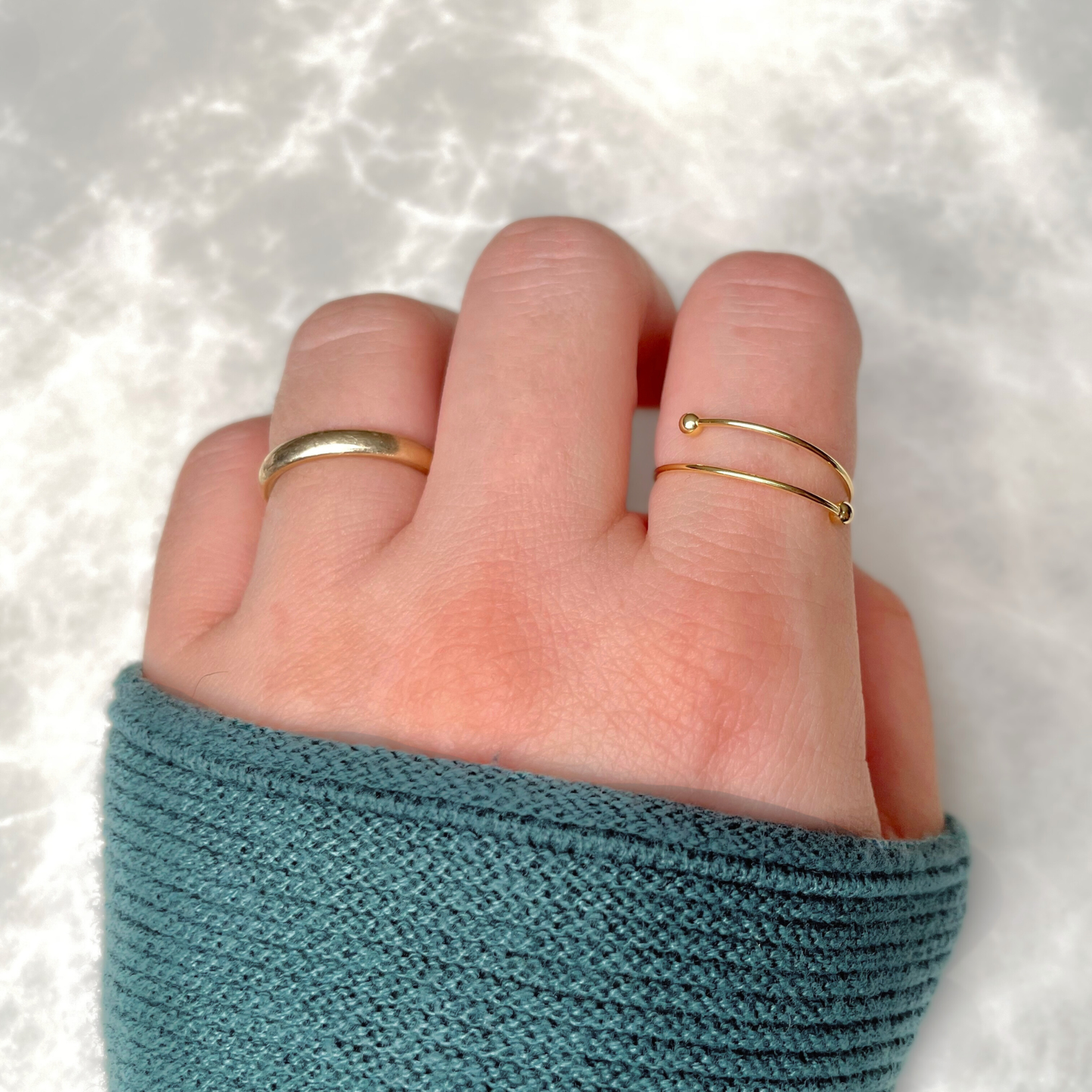 Hand wearing two gold rings, one being the beaded ring on index finger, on a textured white background