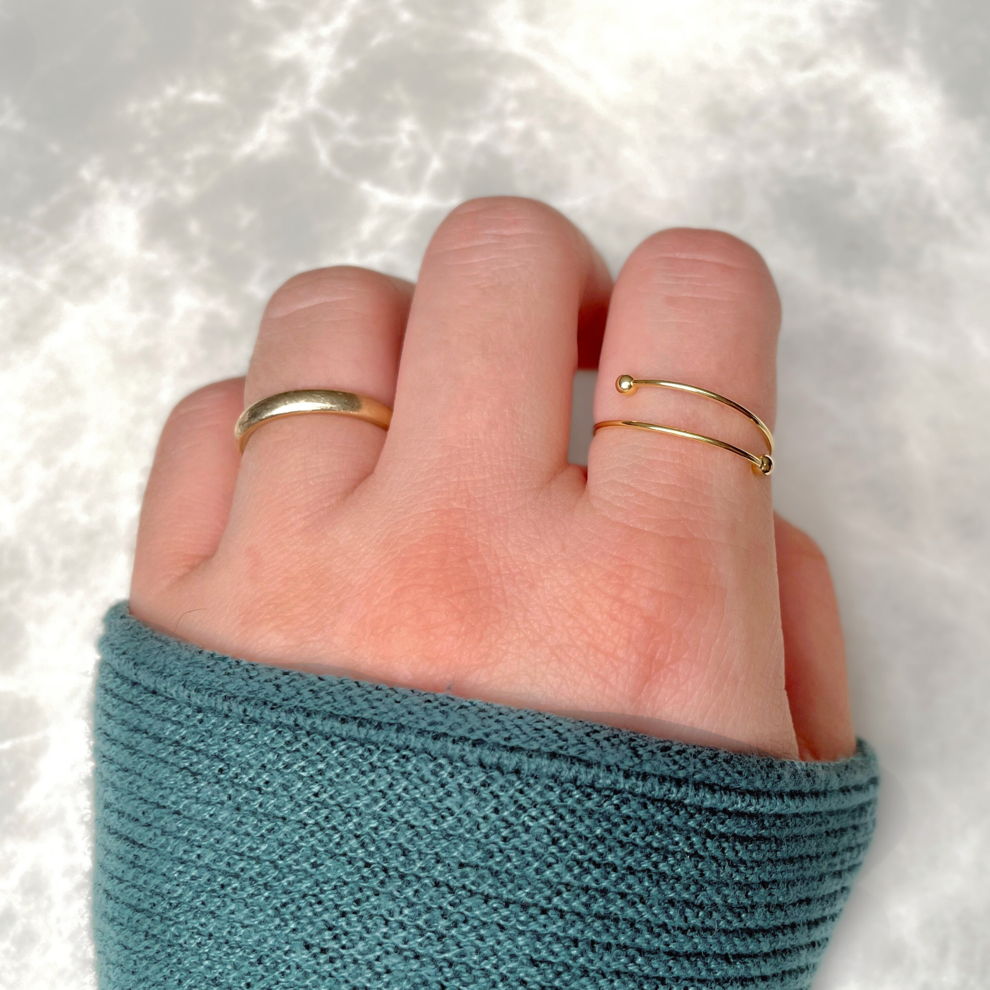 Hand wearing two gold rings, one being the beaded ring on index finger, on a textured white background