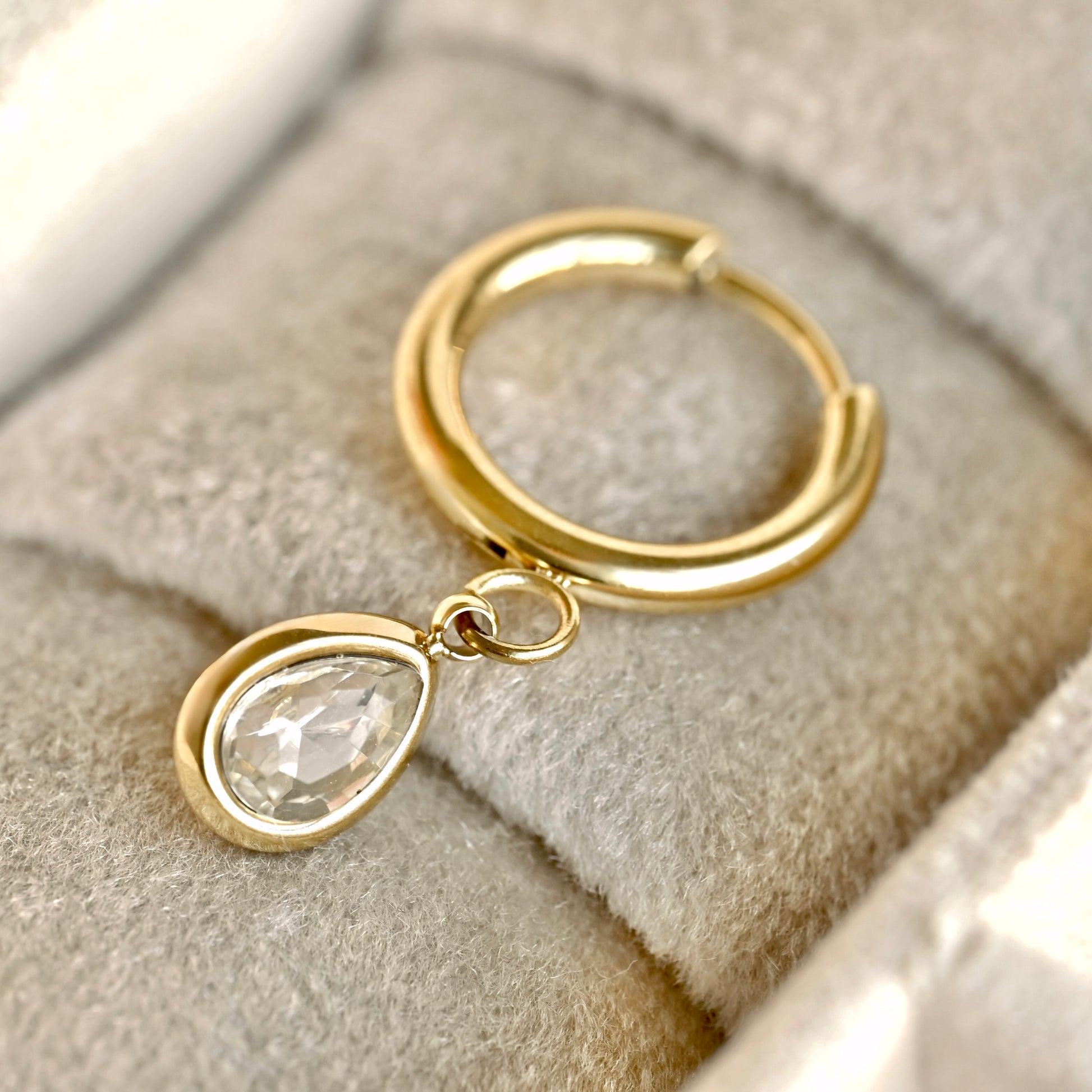 A gold huggie hoop earring with a dangling teardrop-shaped clear faceted gemstone, displayed on a soft gray velvet jewelry box cushion.