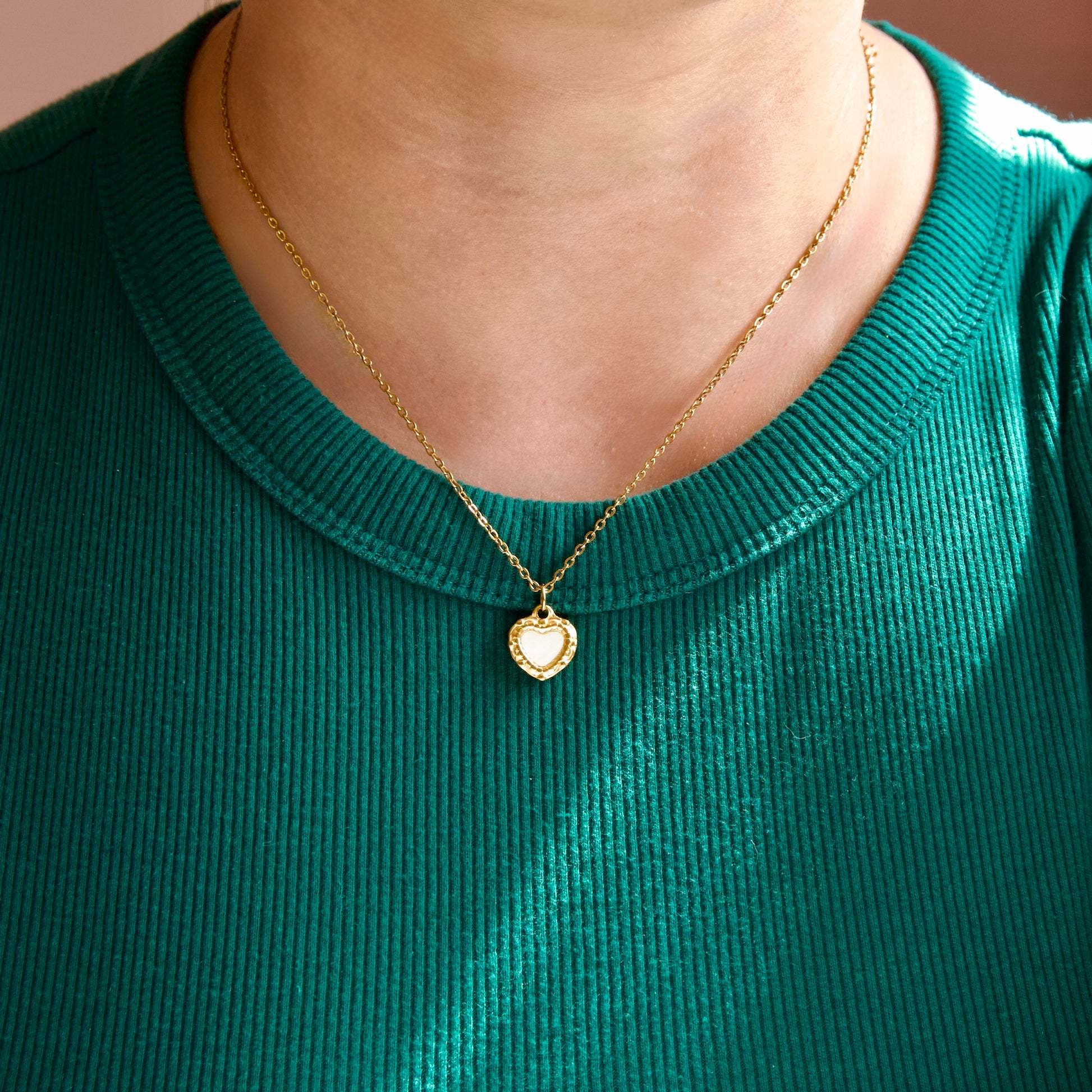 A close-up of a person wearing a delicate gold chain necklace with a small heart pendant, styled with a ribbed green shirt.