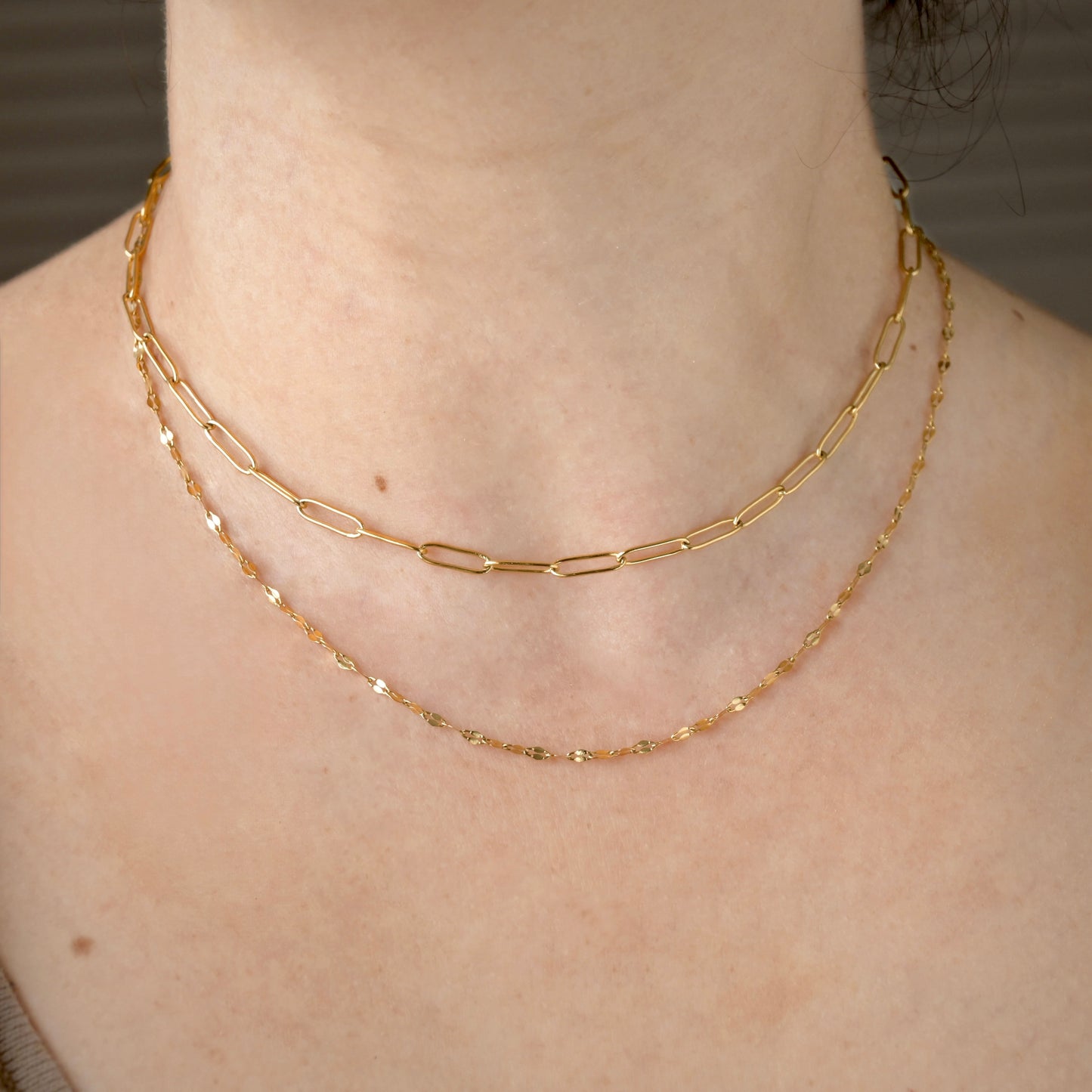 Gold mirror chain necklace layered with a choker length paperclip chain on a person's neck with a neutral background