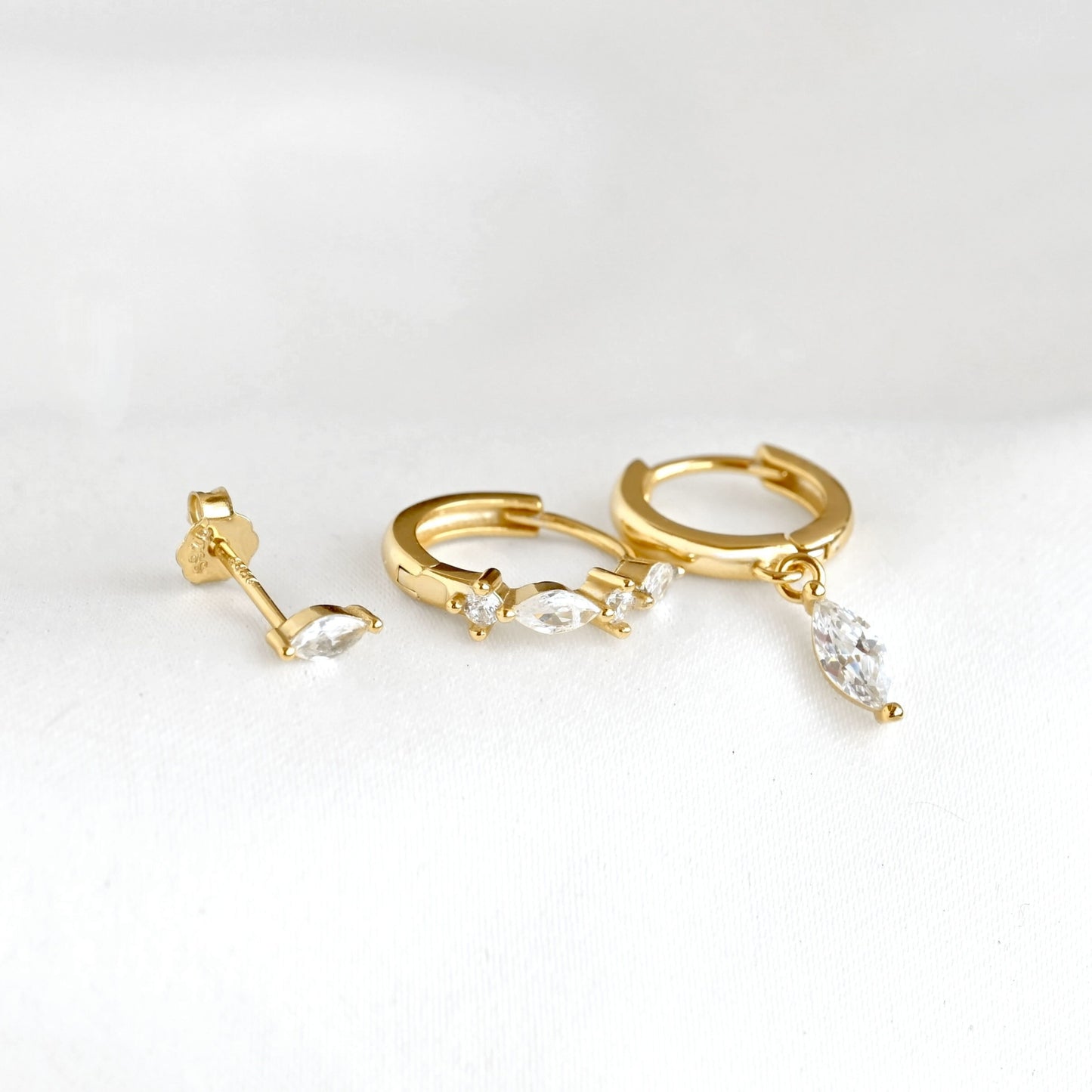 Gold huggie hoop earrings with marquise-cut clear crystals  laid flat to display the dangling design, on a white background.