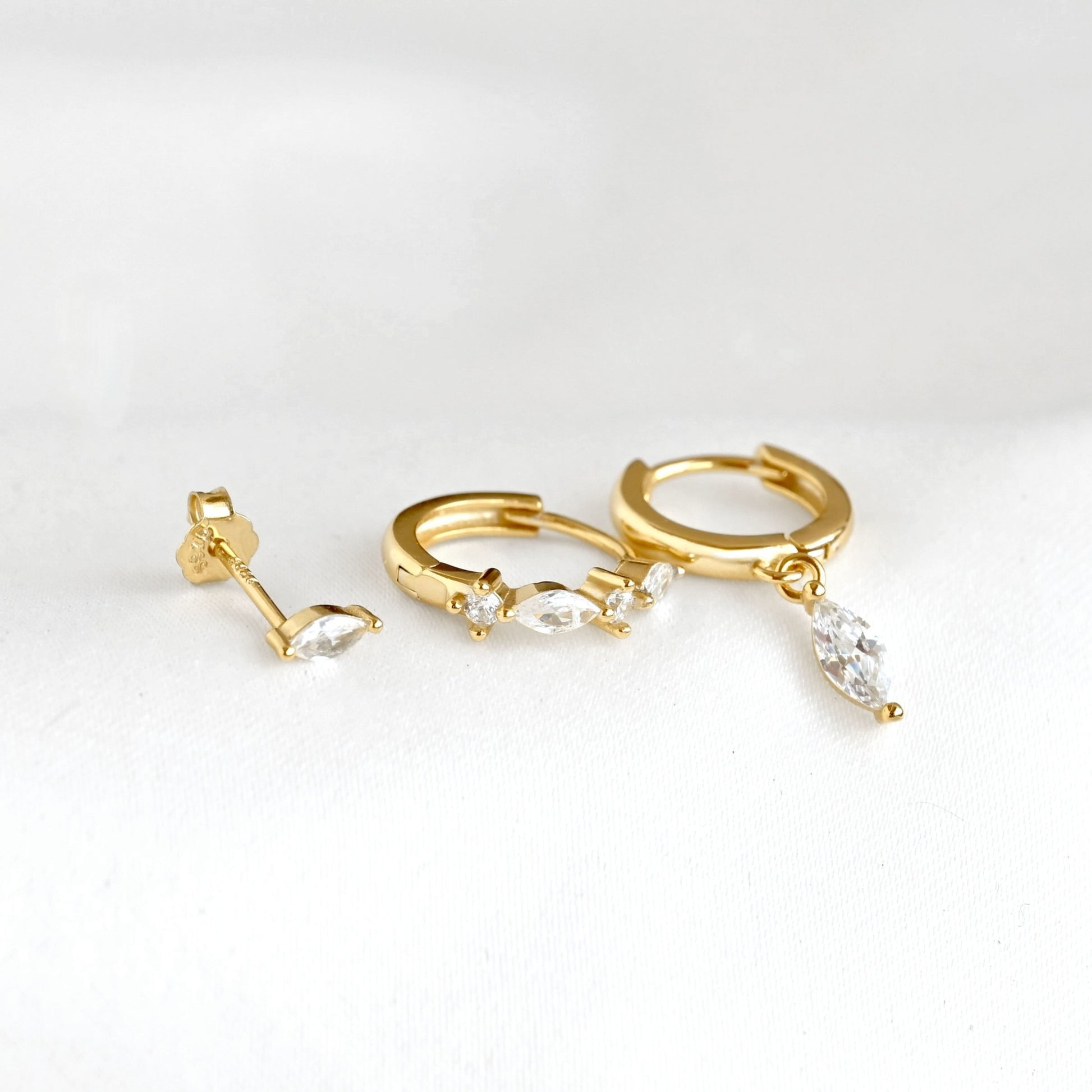 Gold huggie hoop earrings with marquise-cut clear crystals  laid flat to display the dangling design, on a white background.