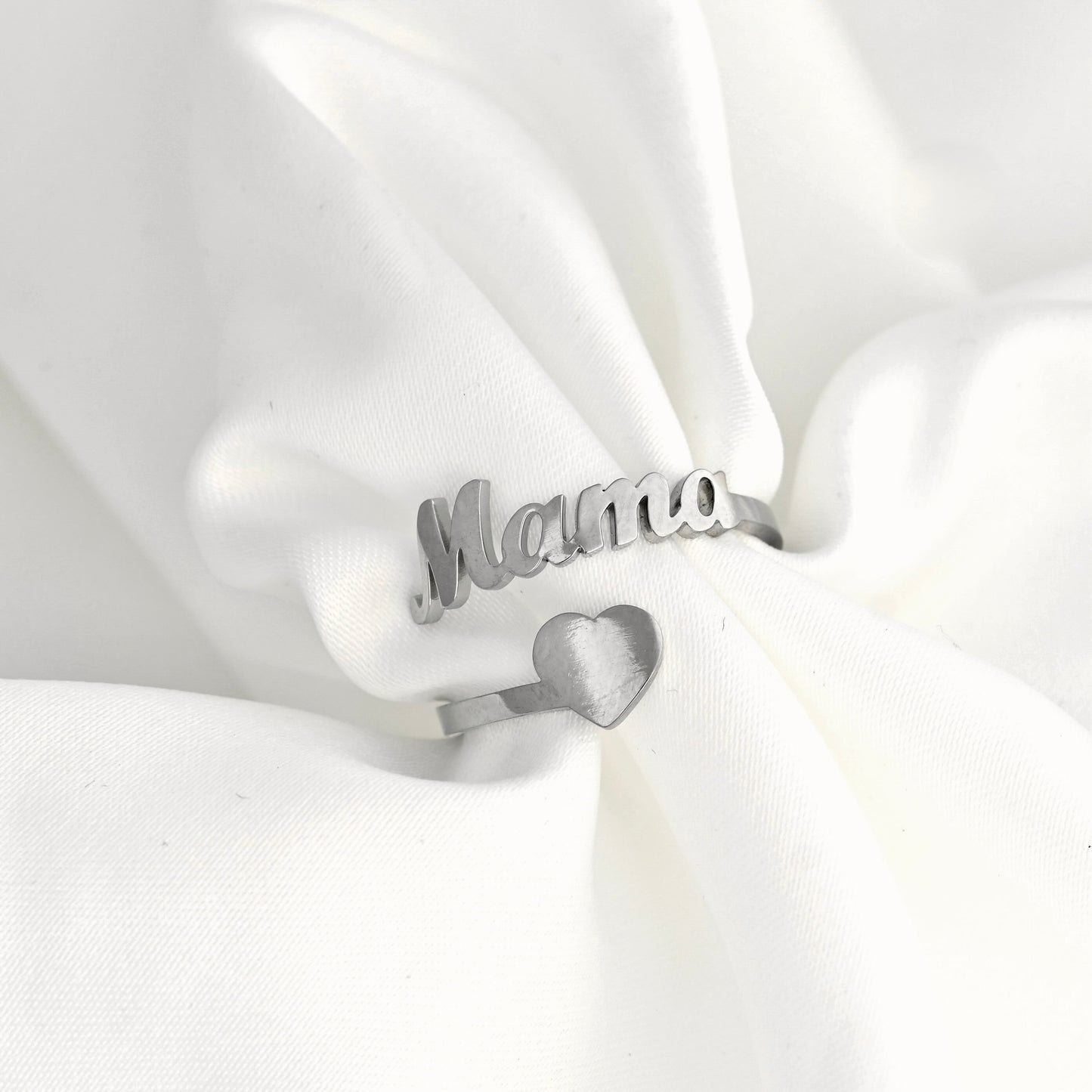A silver ring with the word 'Mama' in cursive script and a small heart charm, displayed on soft white fabric.