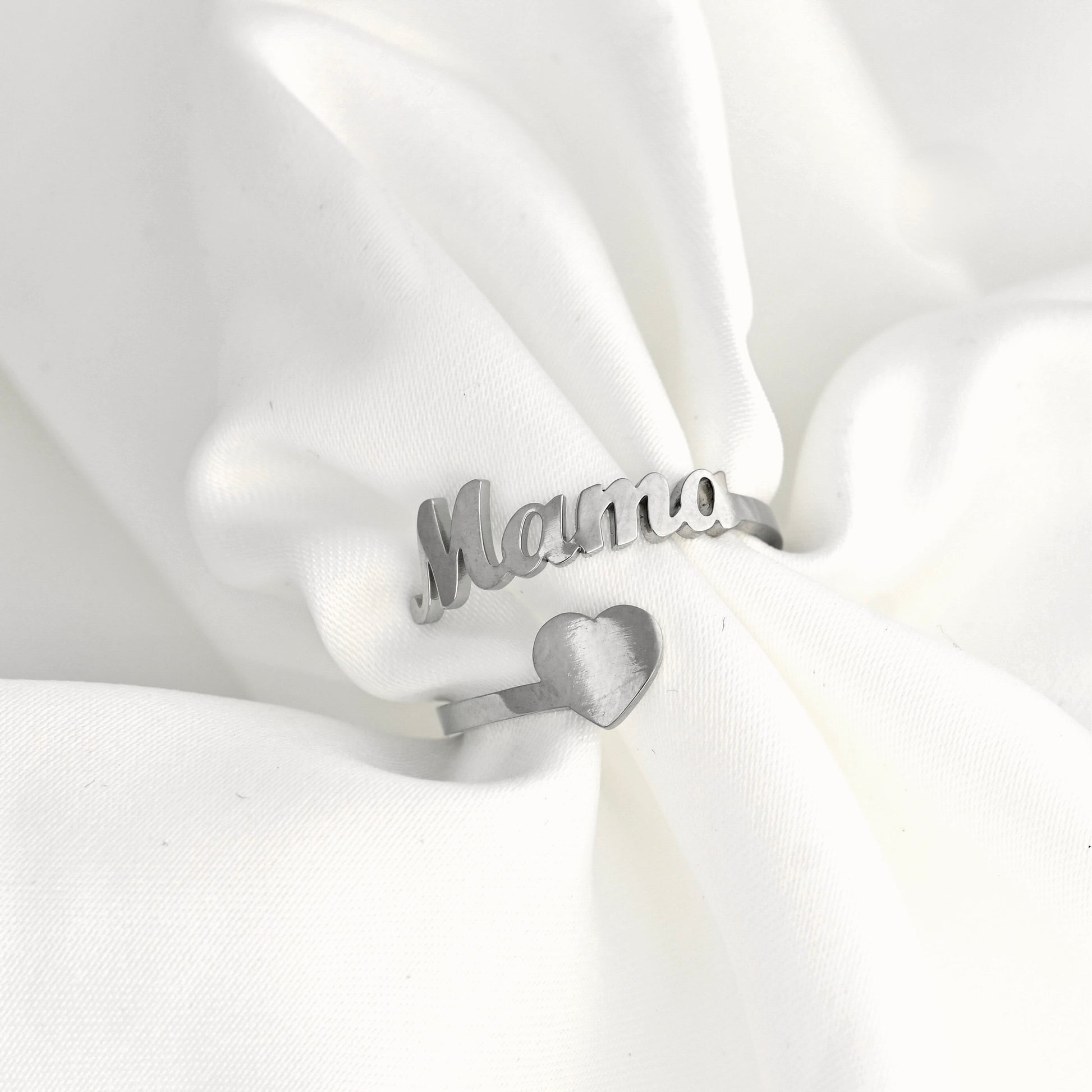 A silver ring with the word 'Mama' in cursive script and a small heart charm, displayed on soft white fabric.