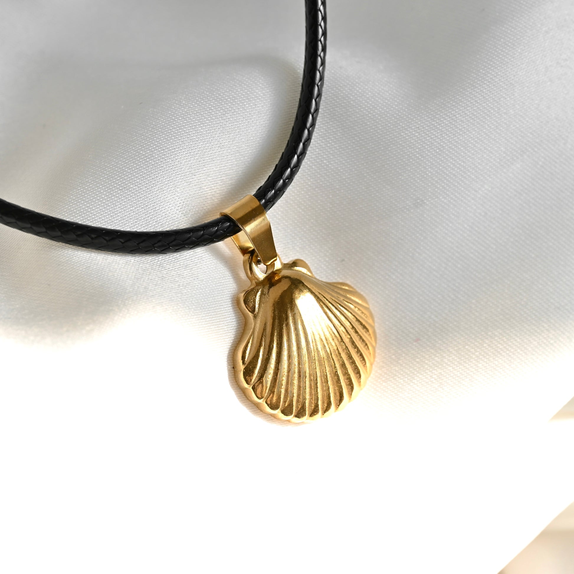A gold-toned shell-shaped pendant necklace on a black cord, displayed on a white fabric background.