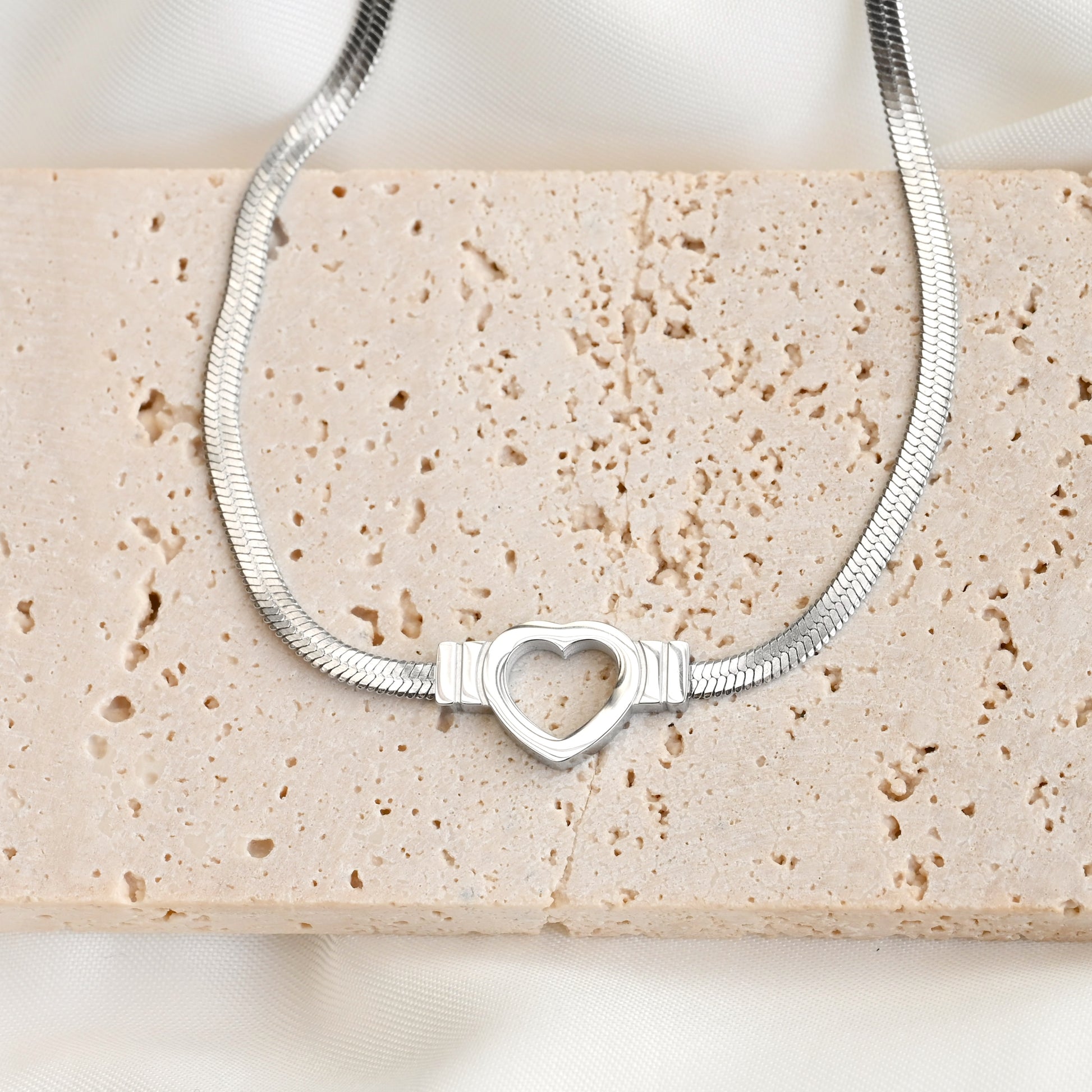 Silver herringbone chain with a heart-shaped center on a textured beige  stone.