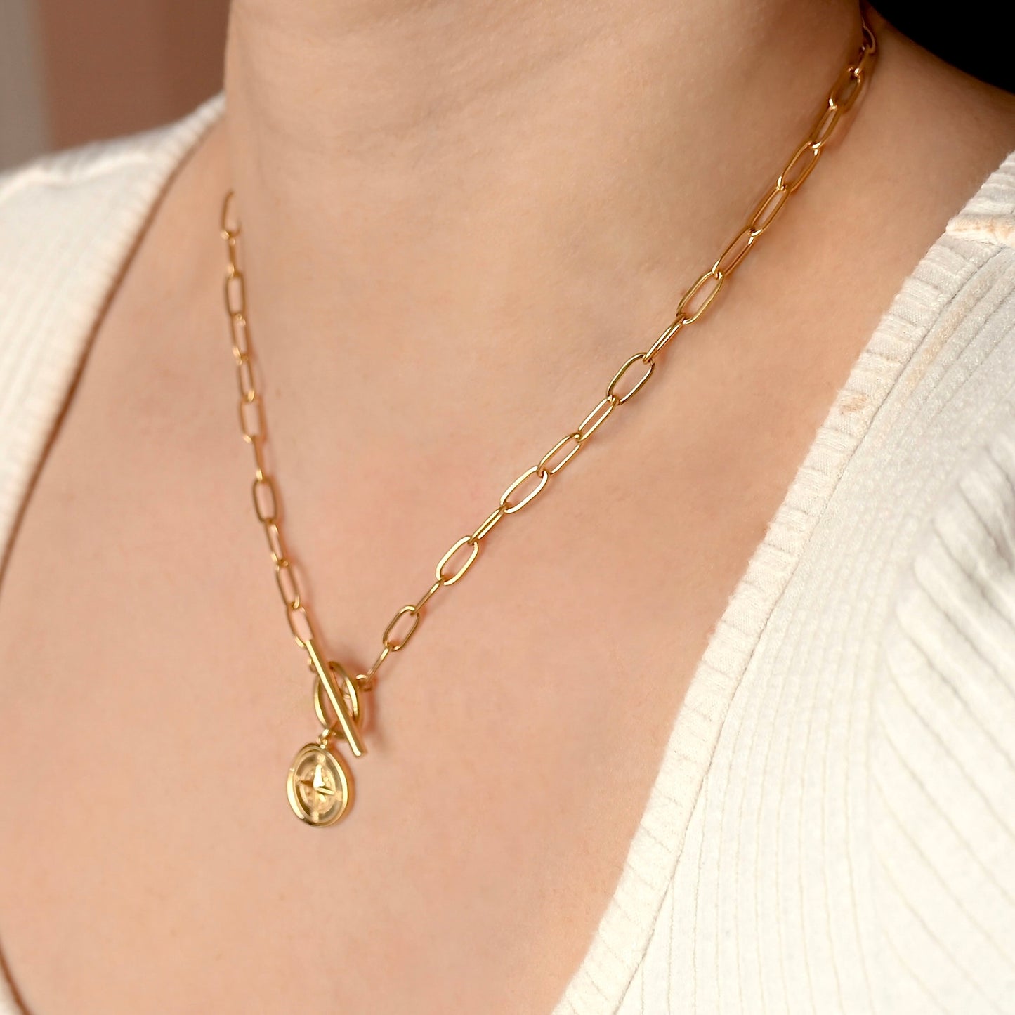 A gold chain necklace with a toggle clasp and a small oval pendant featuring an interlocking double-G style emblem, worn on a model's neck over a white ribbed top.