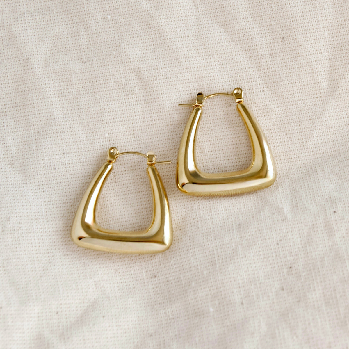 A pair of polished gold-tone geometric hoop earrings with a thick, open triangular design, displayed on a textured off-white linen fabric background.