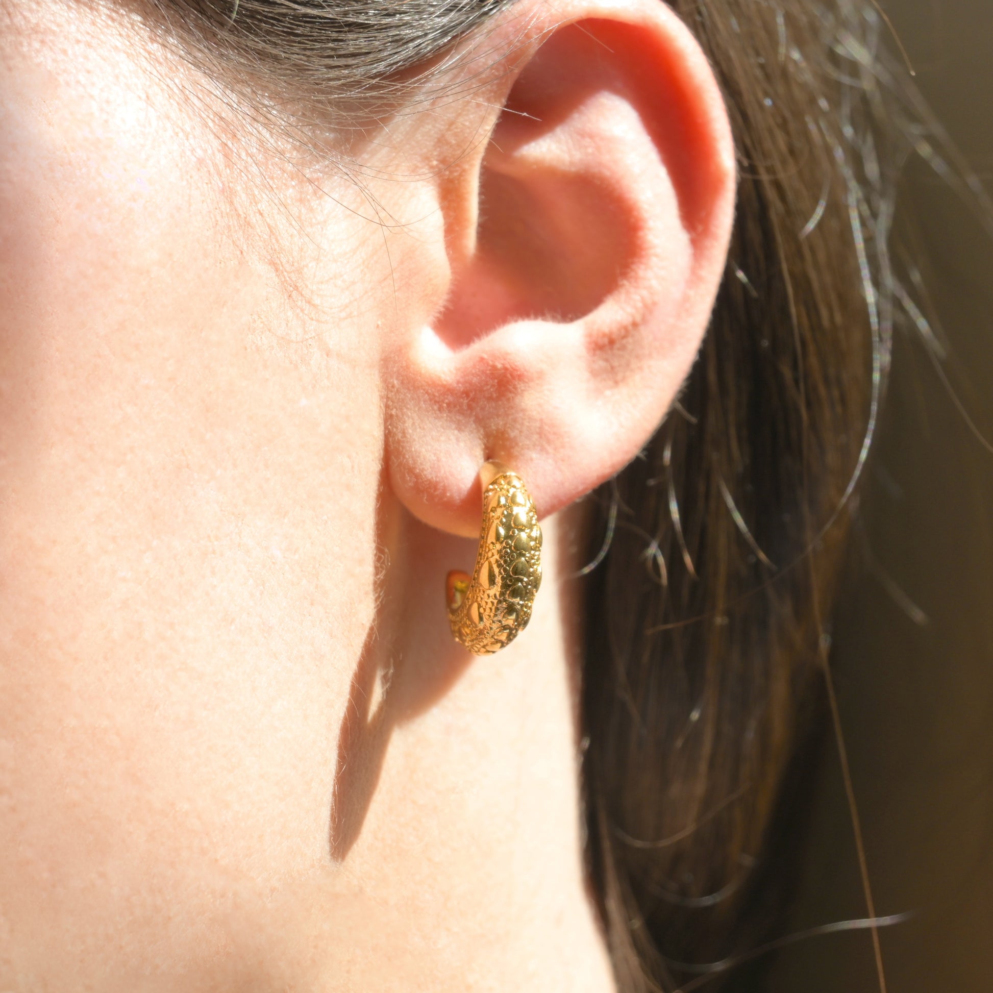 Close-up of an ear wearing a gold C hoop earring  un der direct sunlight