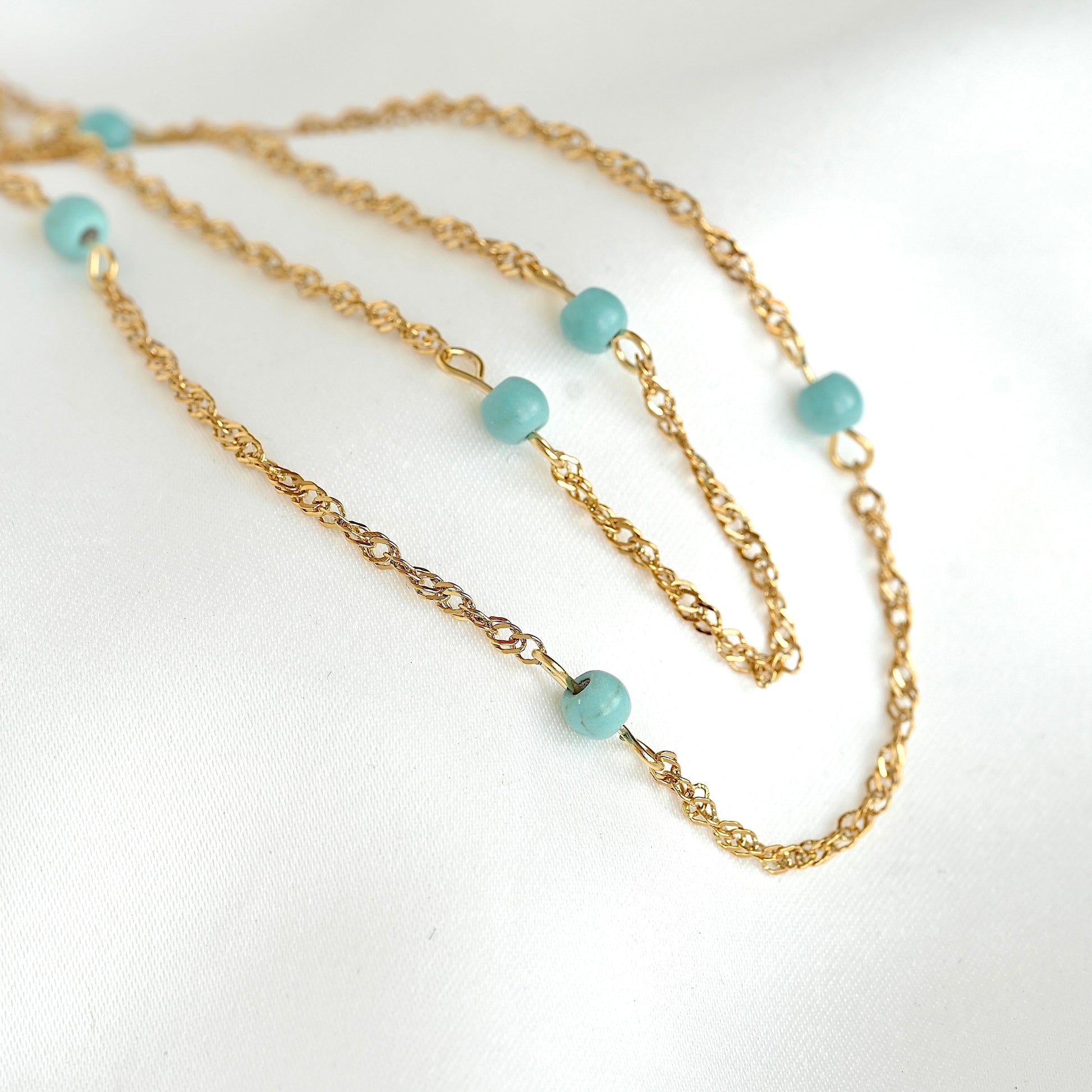 A gold chain necklace with small, round light blue-green beads spaced along the chain, displayed on a soft white fabric background.