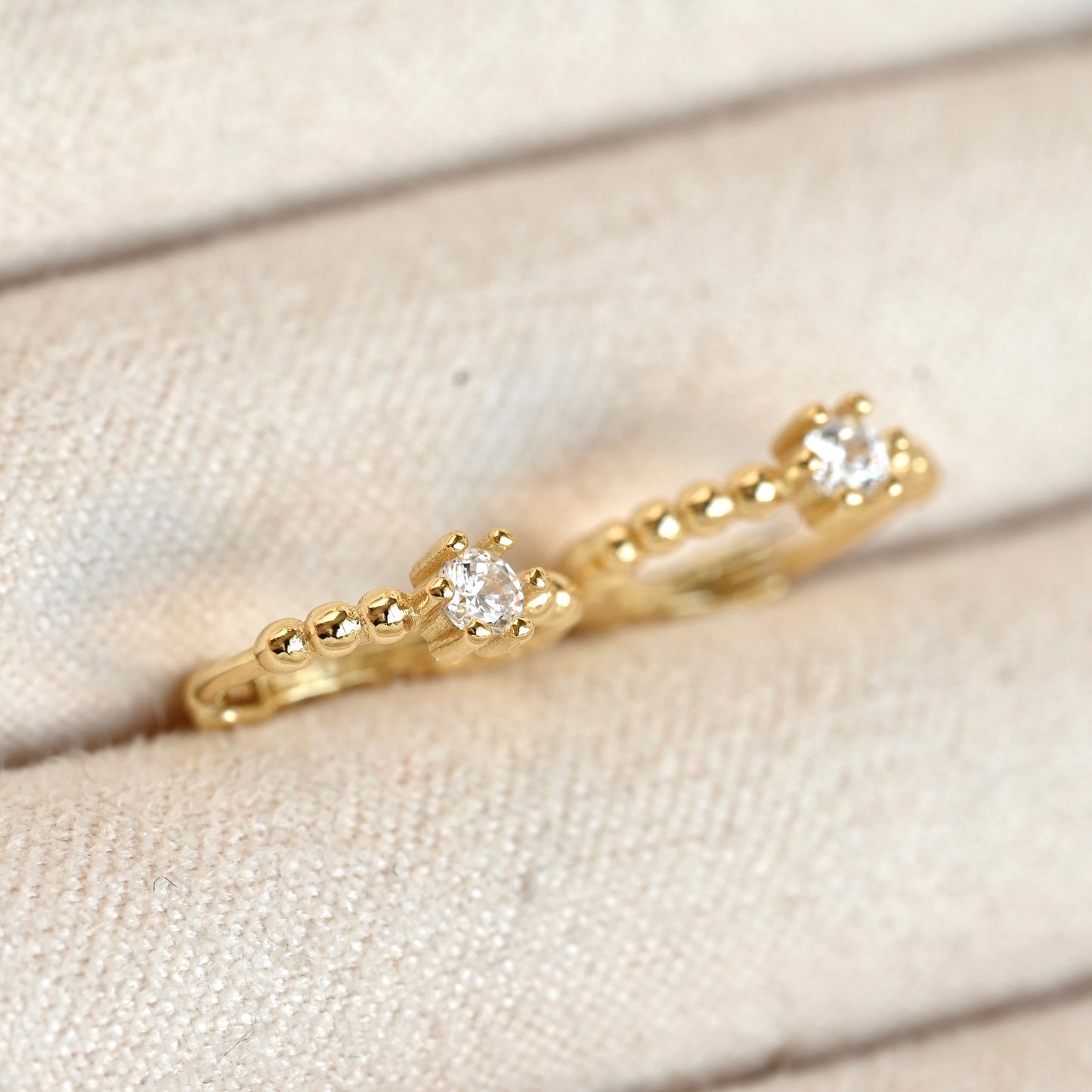 A pair of gold huggie earrings with small clear gemstones, displayed on a textured beige fabric surface.