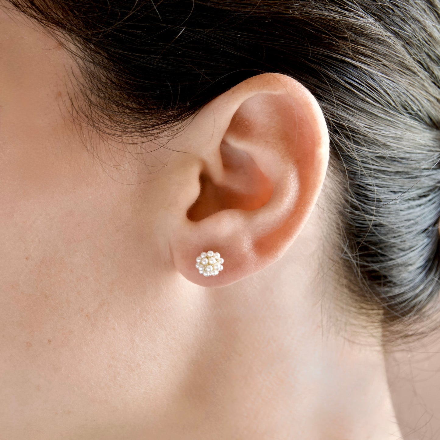 A close-up of a person's ear wearing a small floral stud earring made of milky white round pearls in a gold-tone setting.