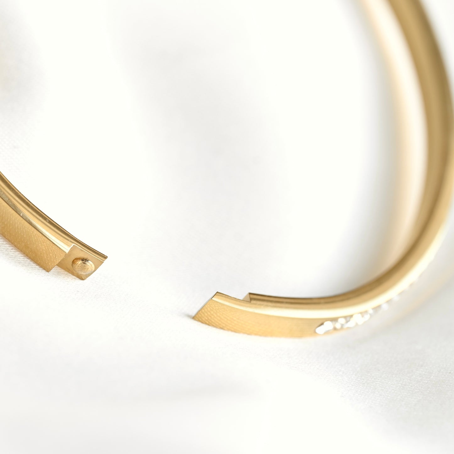 A close-up of a gold-toned open cuff bracelet with a textured finish and a small row of clear stones on one side, photographed on a soft white background.
