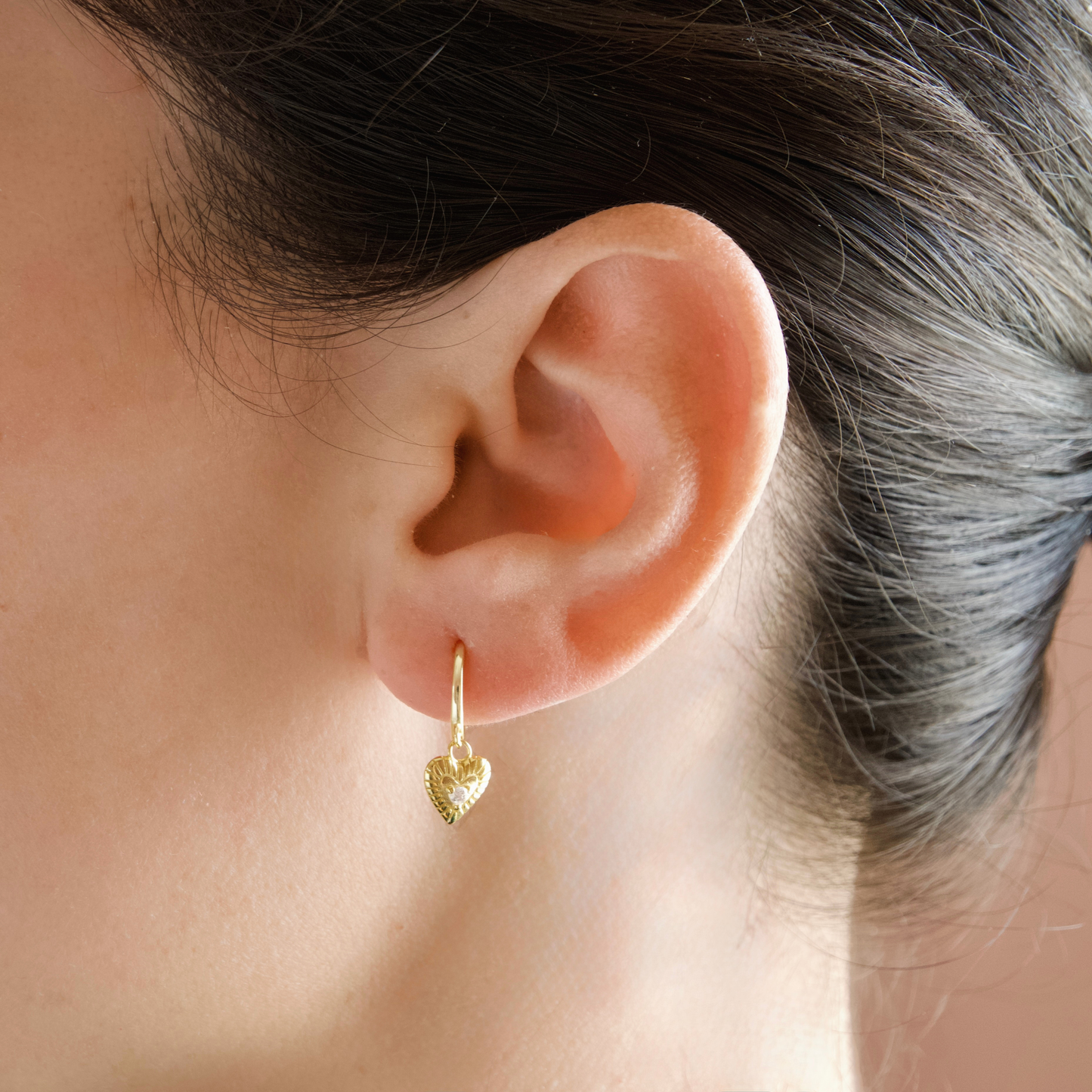 A close-up of a person's ear wearing a small gold huggie hoop earring with a textured heart dangle.