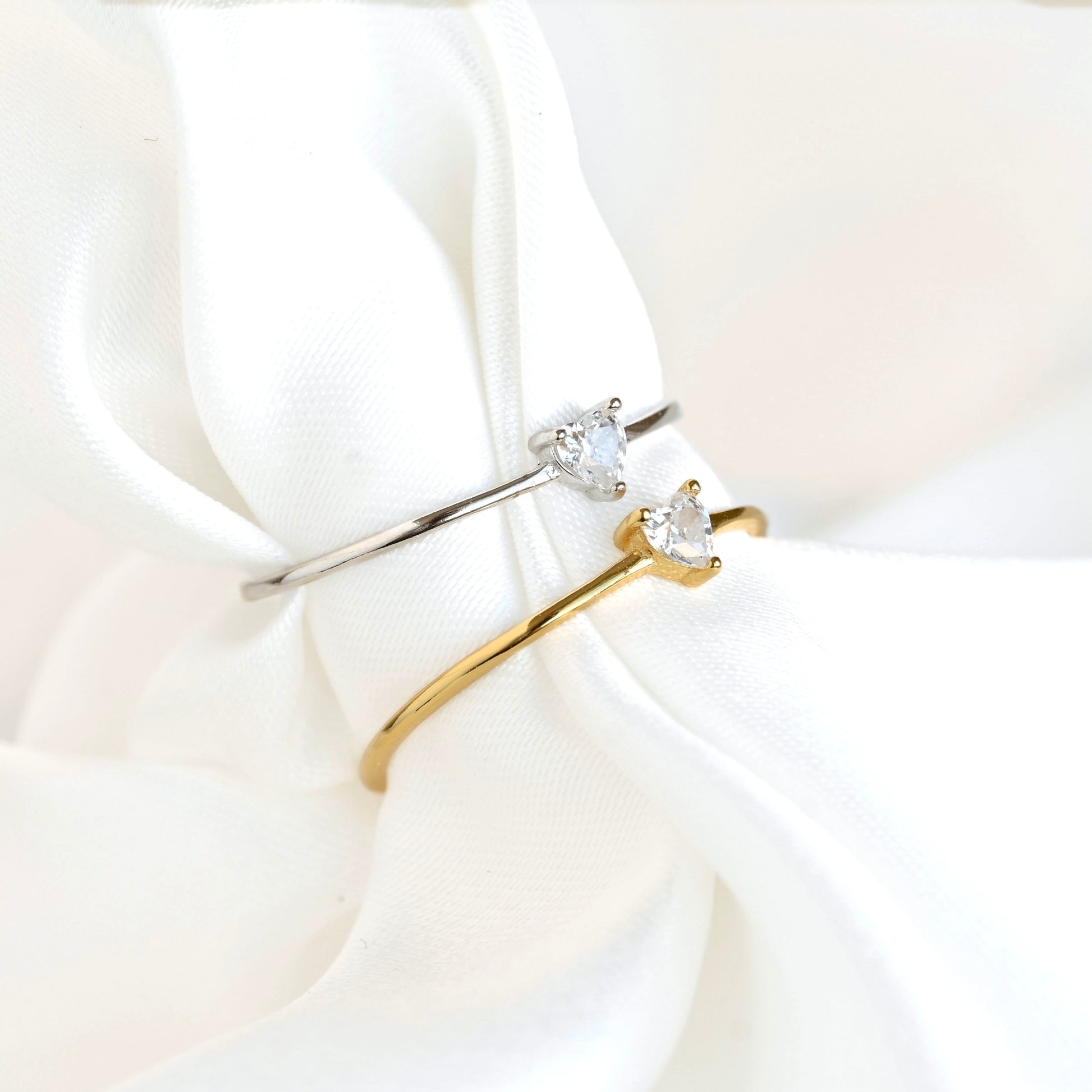 Two slim rings displayed on draped white fabric: one silver band with a small clear heart-shaped stone, and one gold band with a larger clear heart-shaped stone.