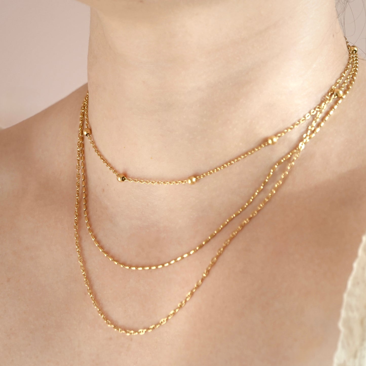 Three gold chains including a satellite chain, oval link chain and mariner chain on a person's neck against a neutral background