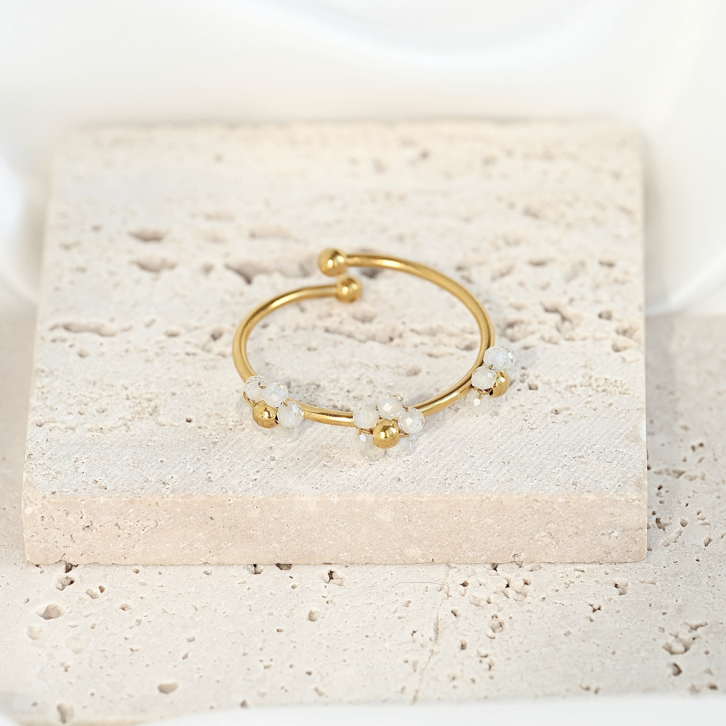 A delicate gold open cuff ring with small clear crystal beads, displayed on a textured beige stone block.