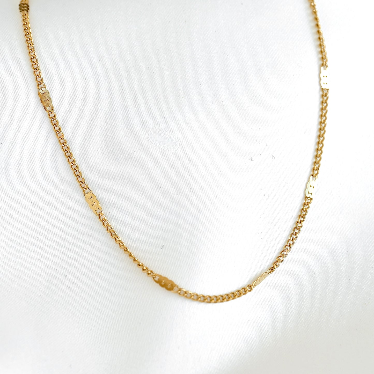 A gold chain necklace with small rectangular sequin beads spaced along the chain, displayed on a white fabric background.