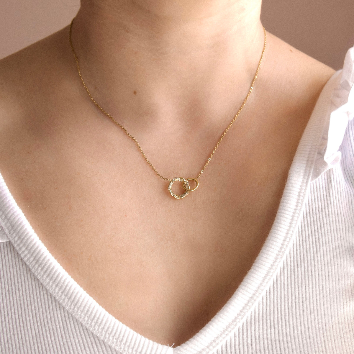 A close-up of a person's neck and chest wearing a delicate gold chain necklace with two interlocking textured rings, paired with a white ribbed V-neck top.