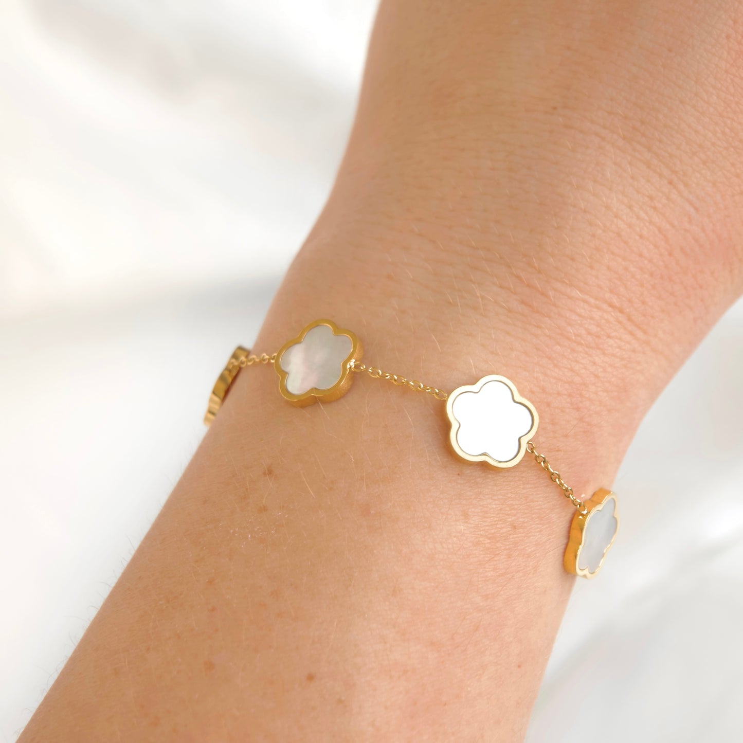 A delicate gold chain bracelet with three white mother-of-pearl clover-shaped charms, shown worn on a person's wrist against a soft white background.