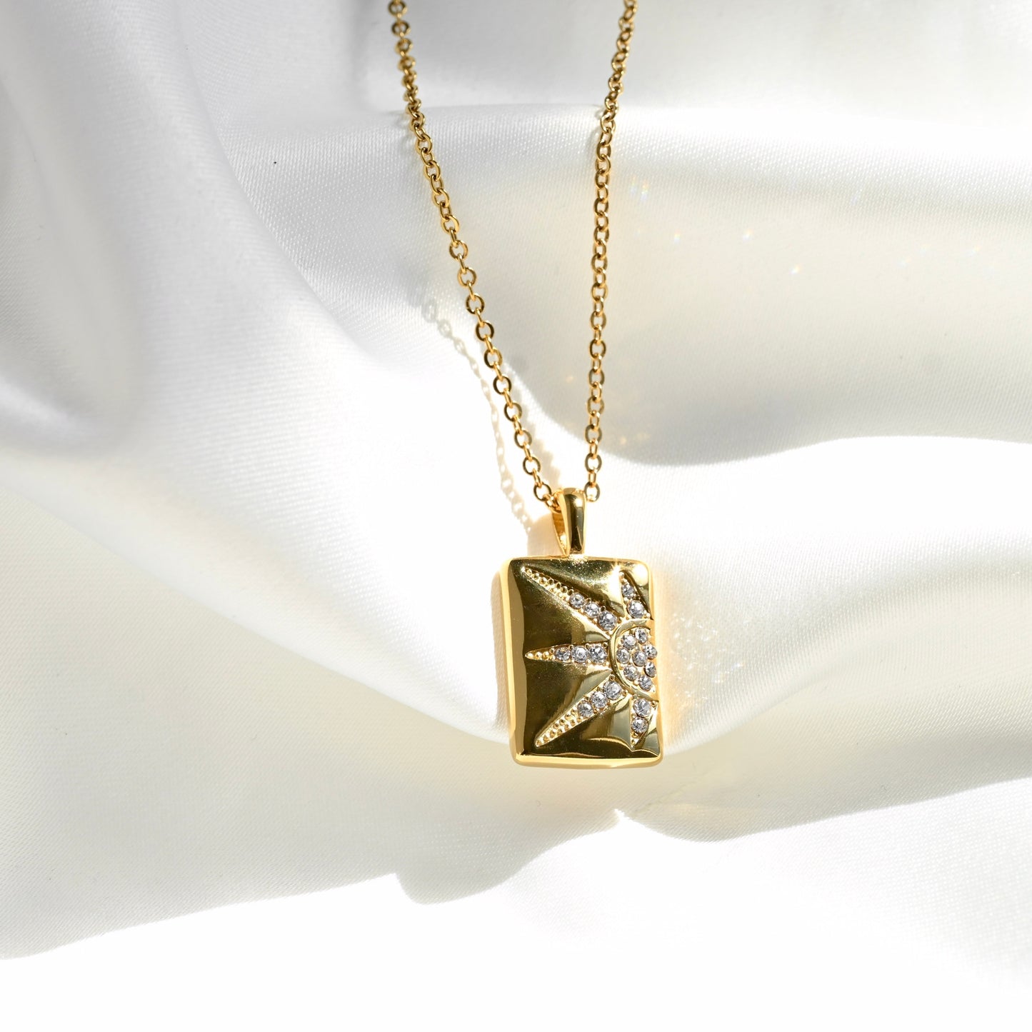 A gold pendant necklace featuring a rectangular gold box with a sunburst design paved with small clear stones, displayed on a white satin fabric background under direct sunlight.