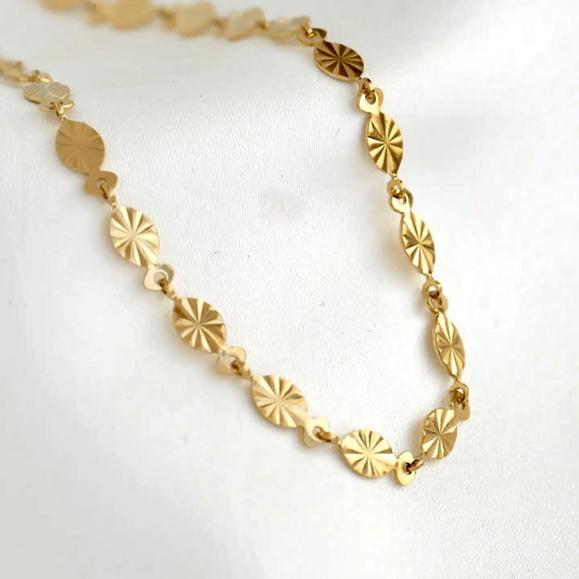 A gold-toned link necklace featuring oval disc charms with a sunburst or starburst textured pattern, displayed on a soft white fabric background.