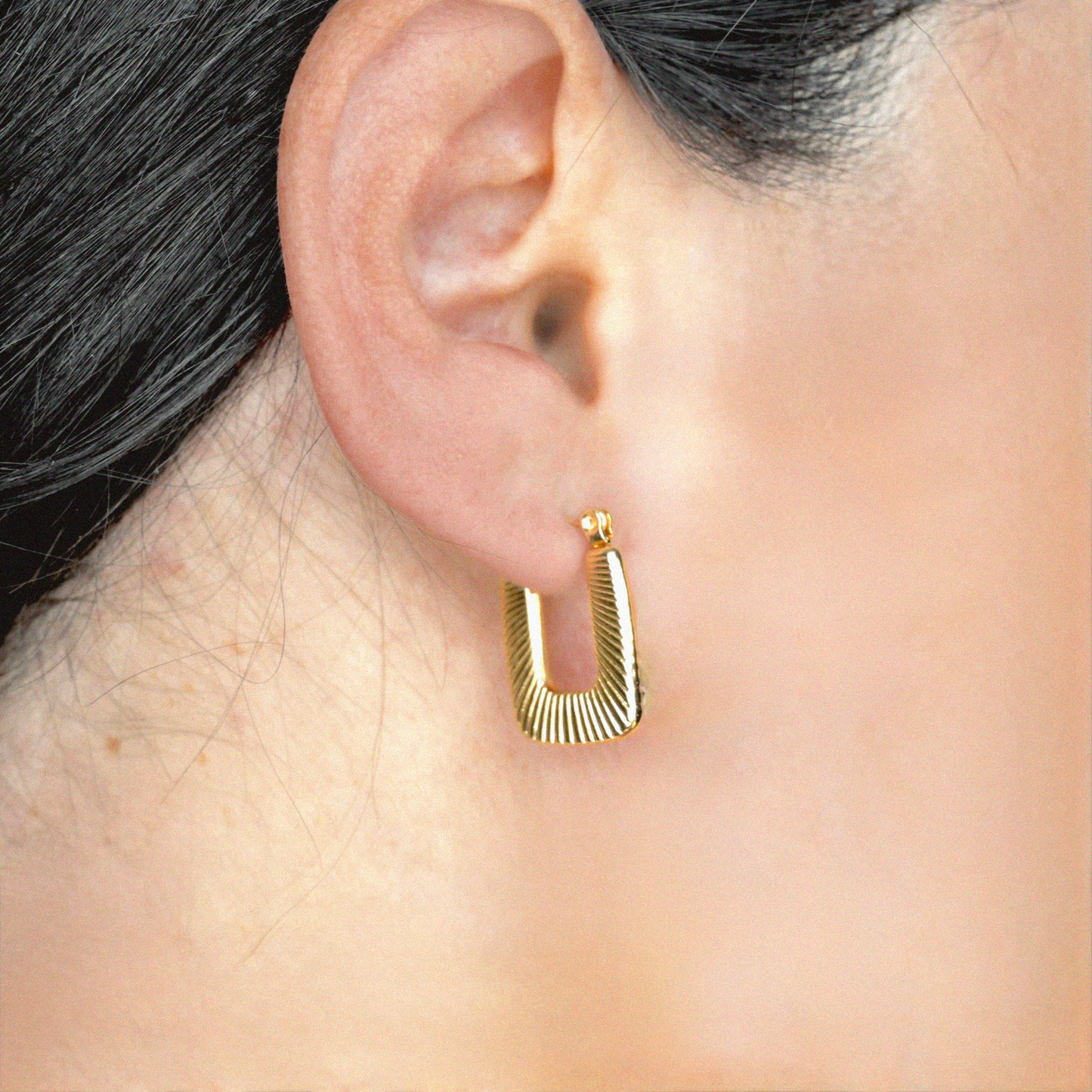 Gold geometric hoop earring worn by a person with dark hair.