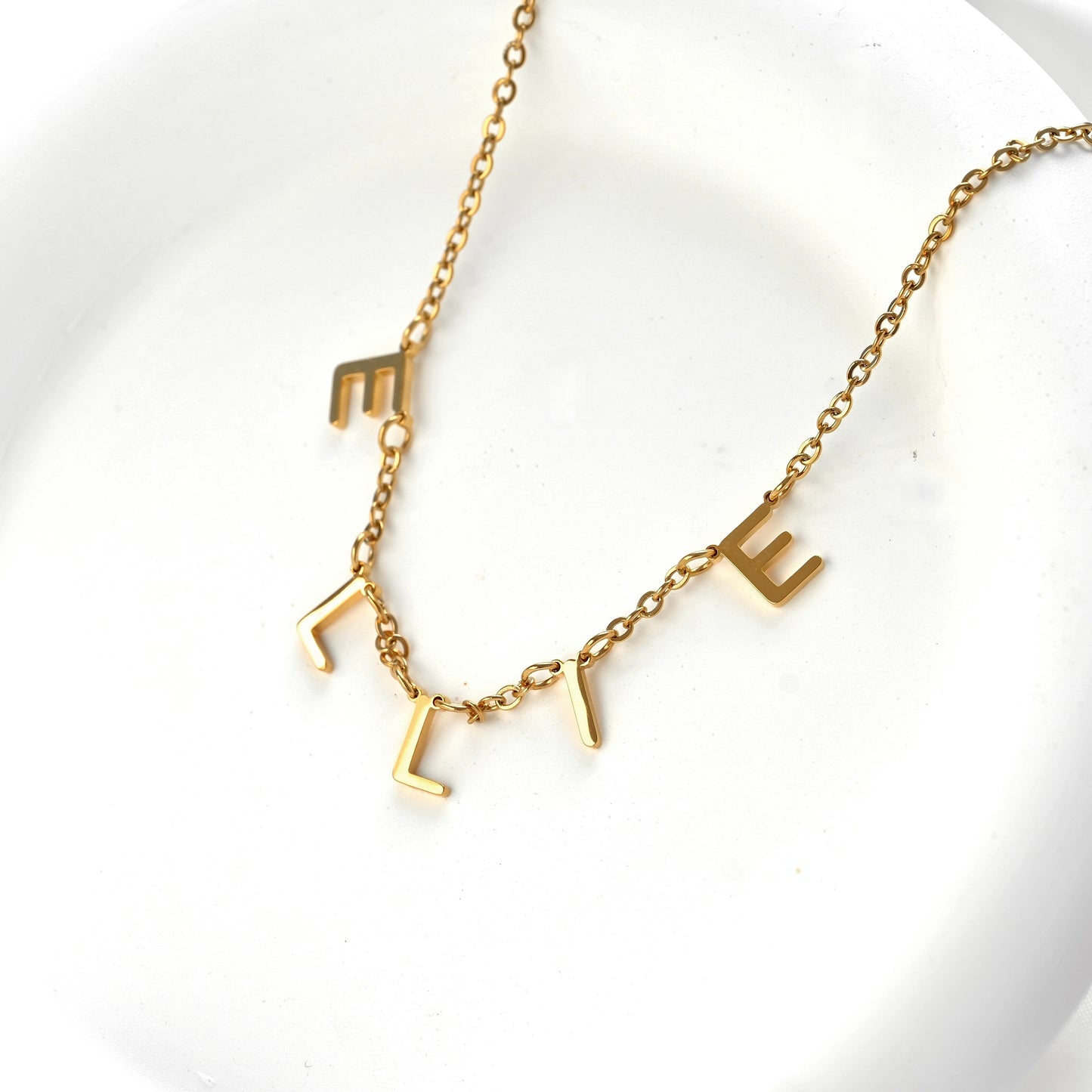 A gold initial necklace spelling 'ELLIE' with letter-shaped charms on a delicate chain, displayed on a white textured surface.